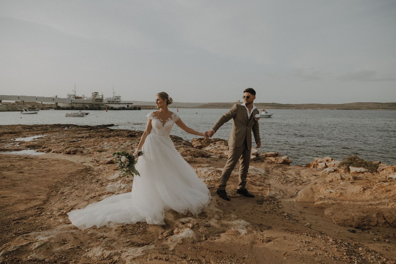 Wedding couple holding hands while standing on coastal rocks during golden hour.