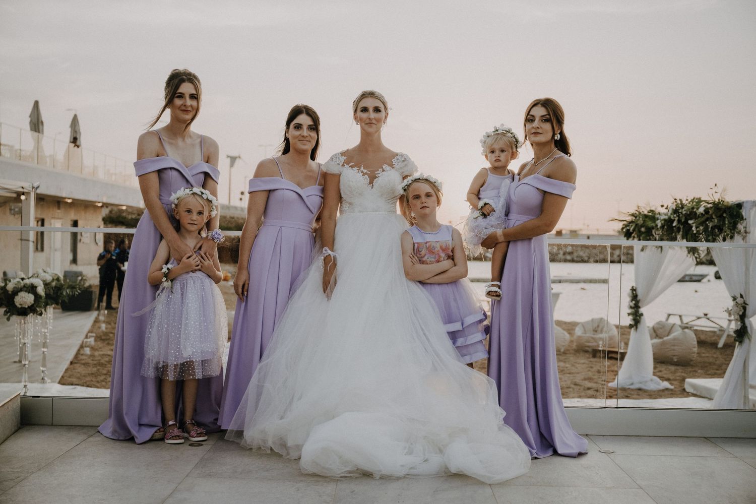 Wedding party lined up in lavender bridesmaid dresses with bride in white gown at outdoor venue.