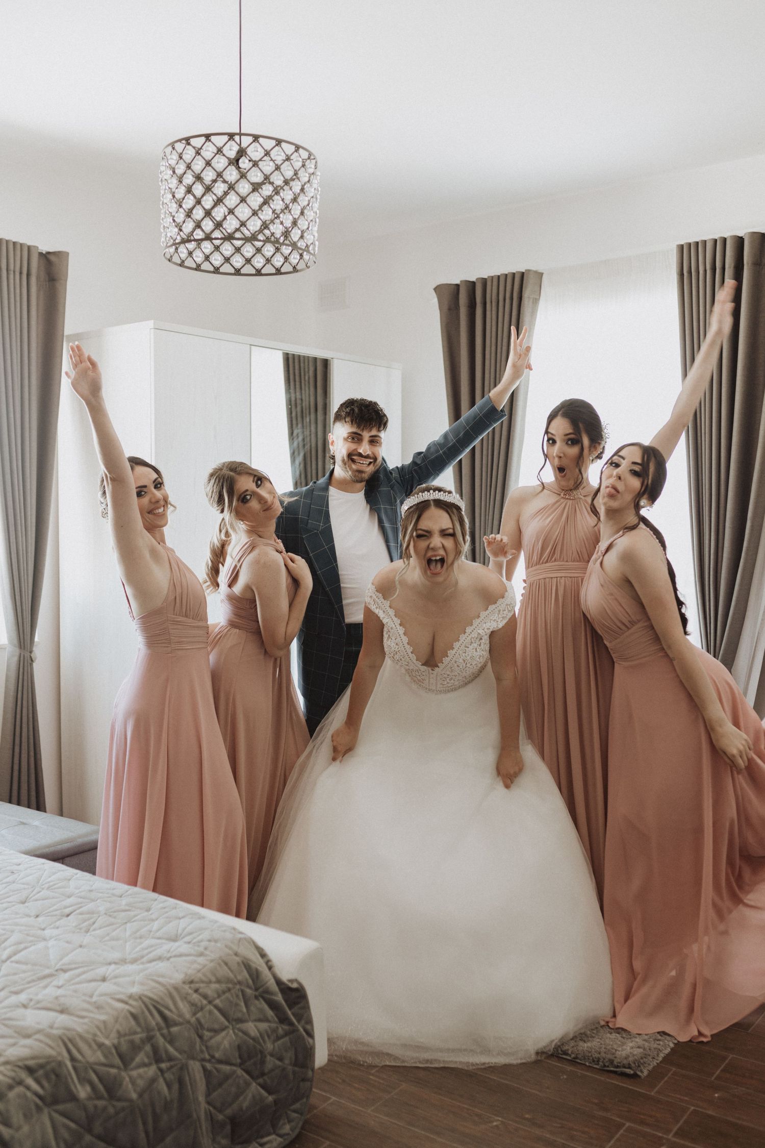 Bridesmaids in blush dresses celebrate joyfully with the bride in her white gown against neutral curtains.