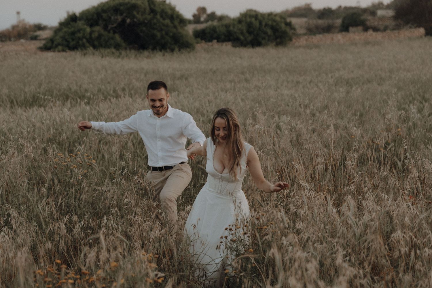 A joyful couple runs through a golden field, the bride in a white lace gown and groom in a white shirt, laughing together.
