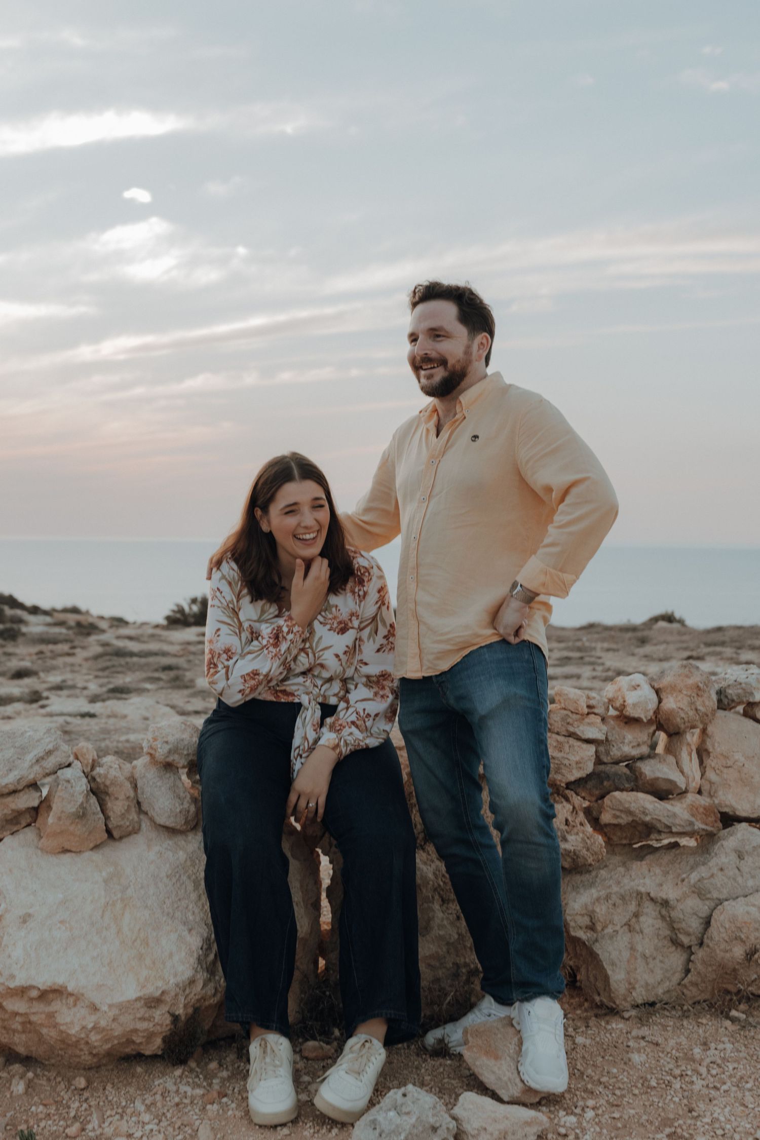 A couple sits casually on rocky terrain during a cloudy evening photoshoot.