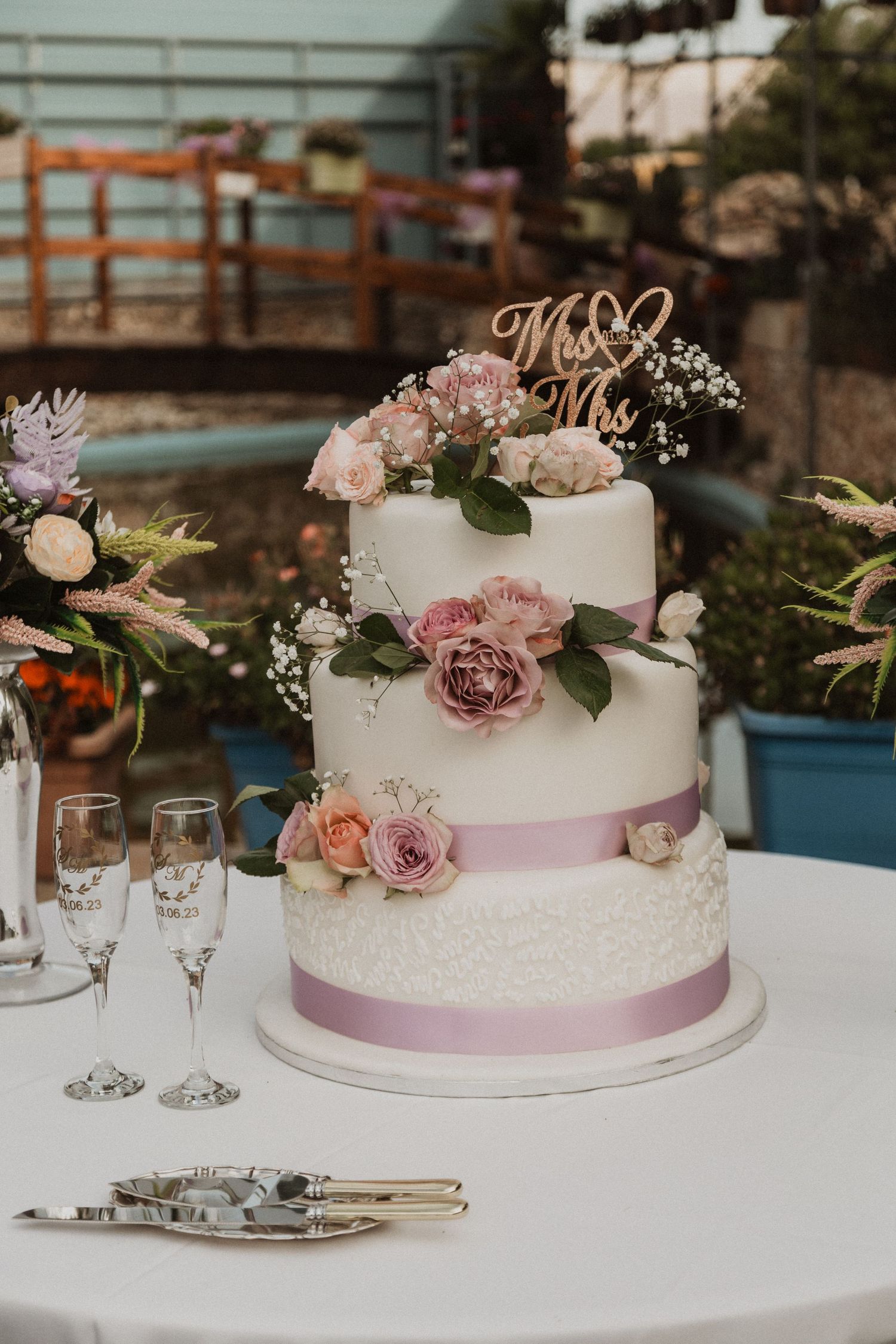 Three-tiered white wedding cake decorated with pink roses and purple ribbon.