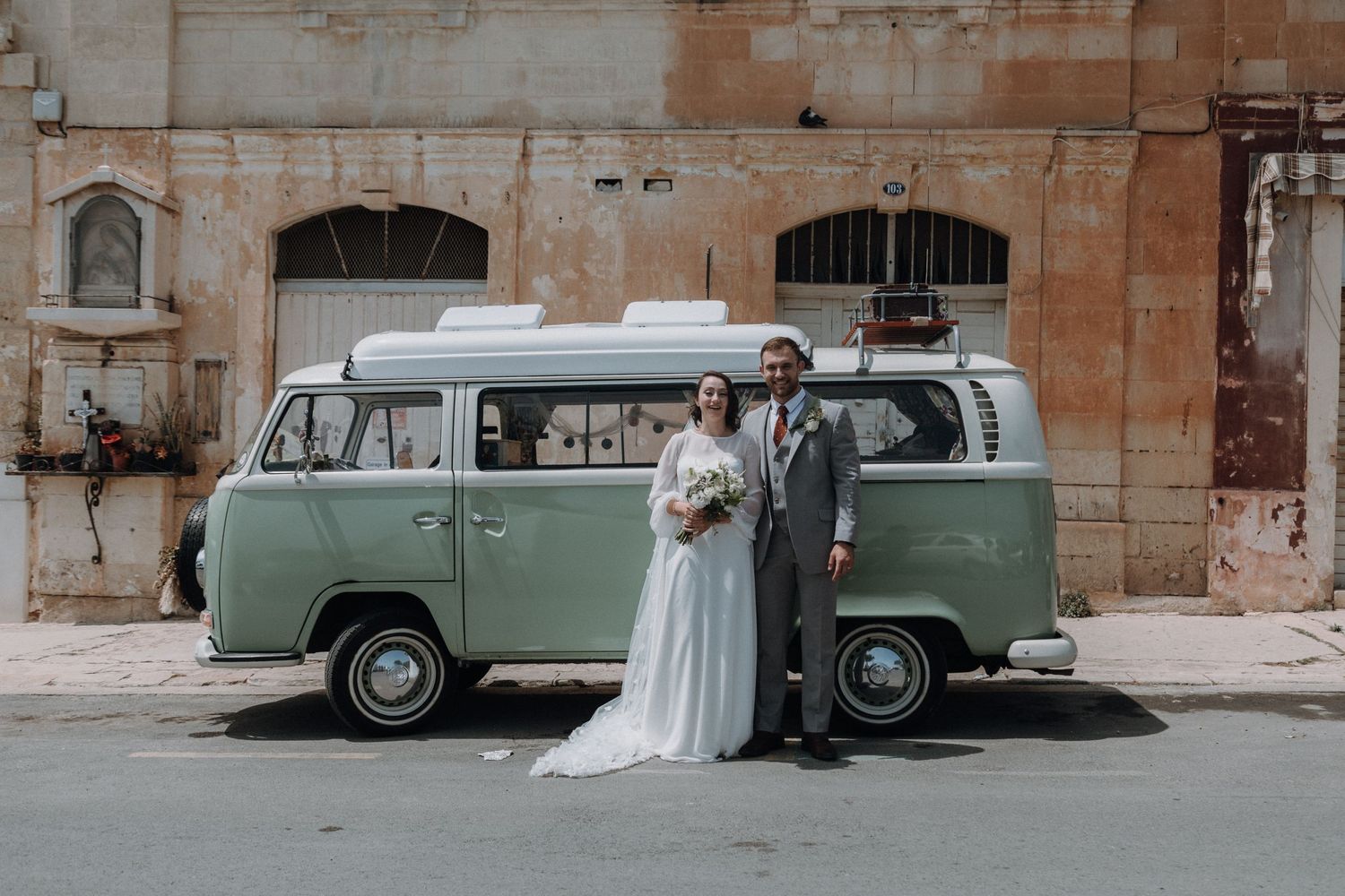 A vintage mint green car parked outside stone buildings with decorative floral arrangements.