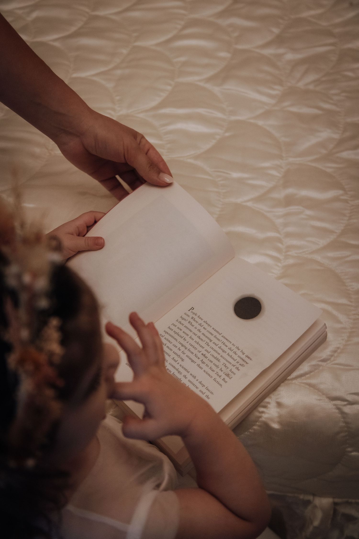 Hands hold open a book on a textured white bedspread, creating an intimate reading moment.
