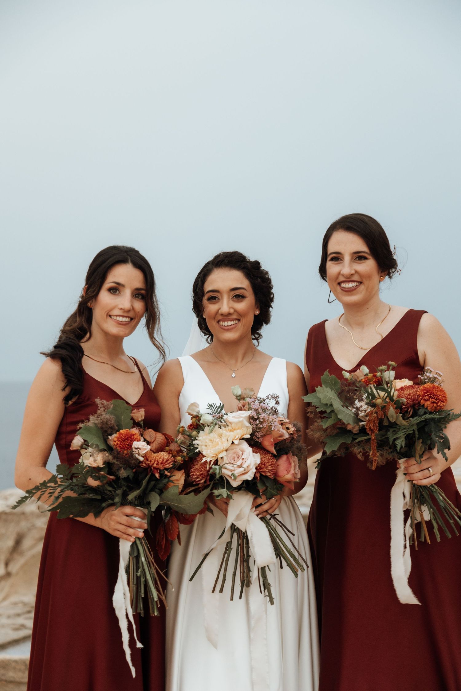 Three women in burgundy and white gowns hold beautiful autumn-toned bouquets against a coastal backdrop.