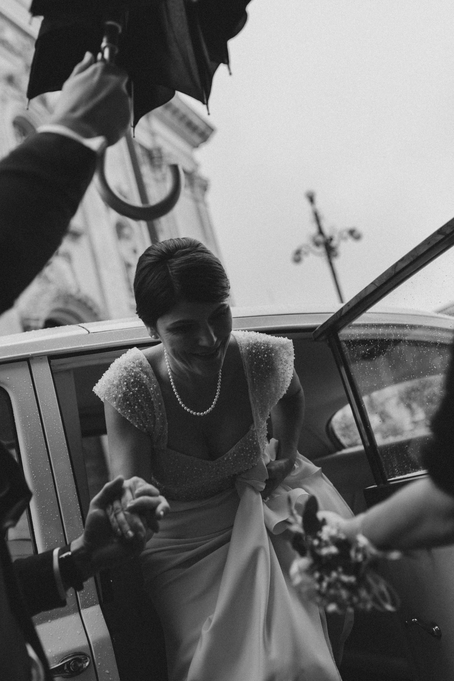 Monochrome photo of a bride in a white dress getting into a classic car.