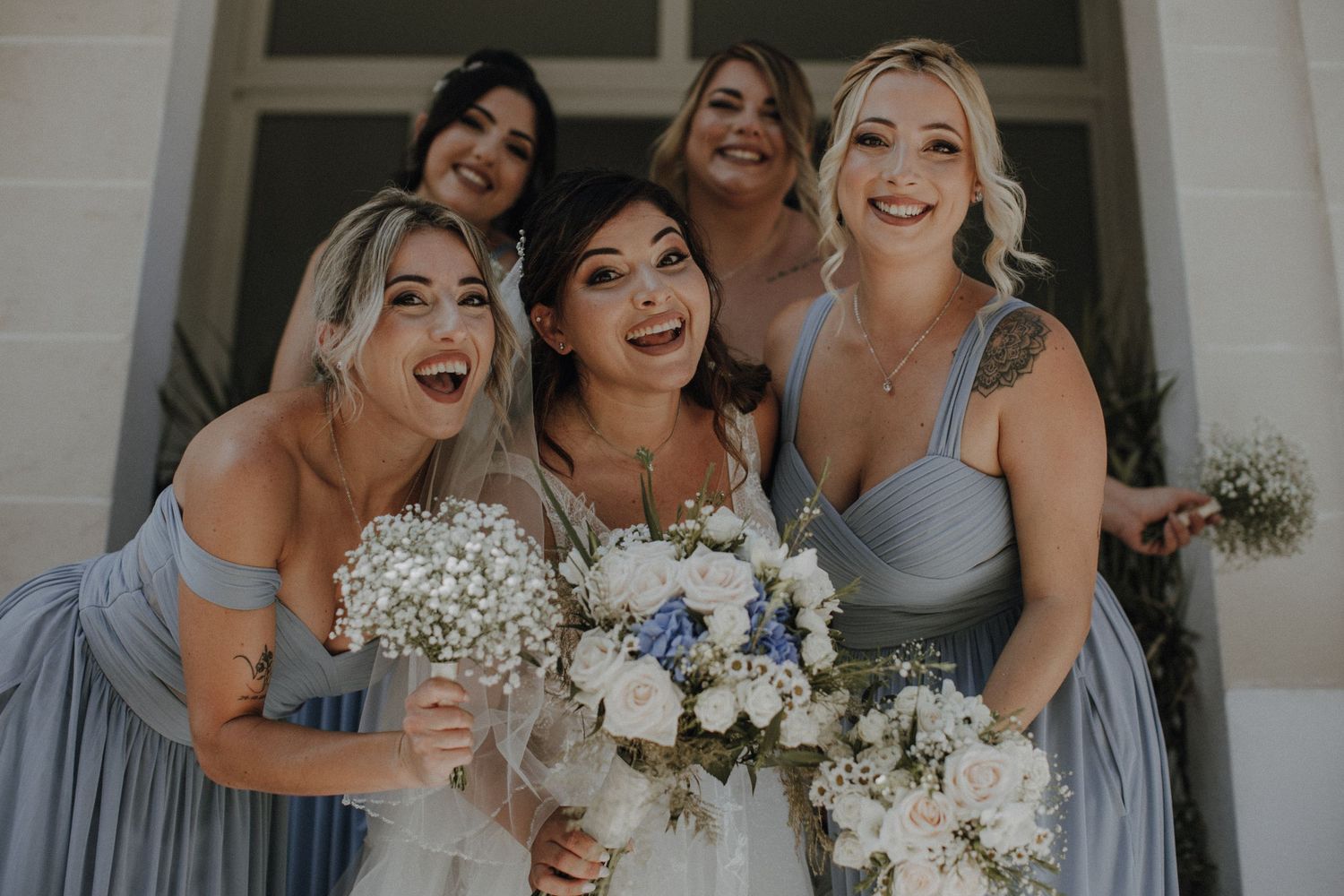 Wedding party smiles with joy while holding white and blue floral bouquets.