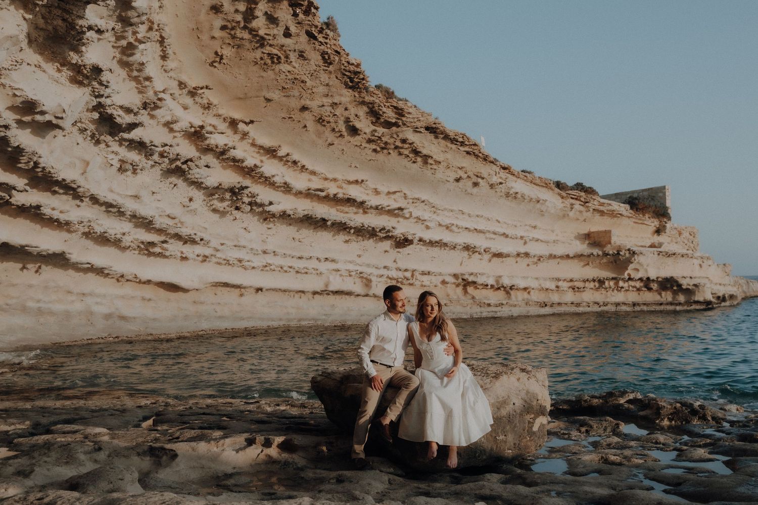 Couple sitting on rocks by the sea with dramatic white limestone cliffs in the background.