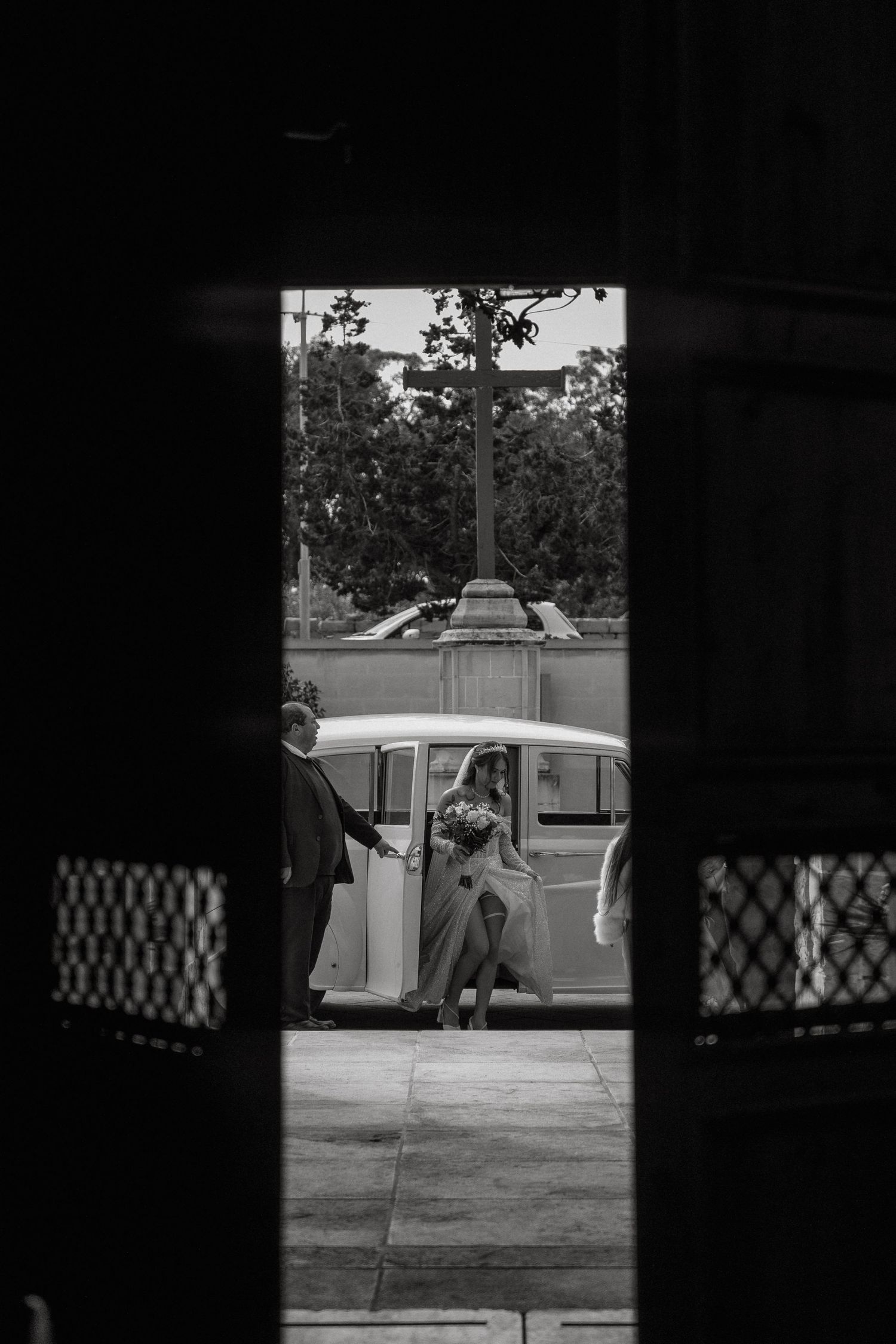 Vintage black and white photo of bride entering classic car through church doorway.