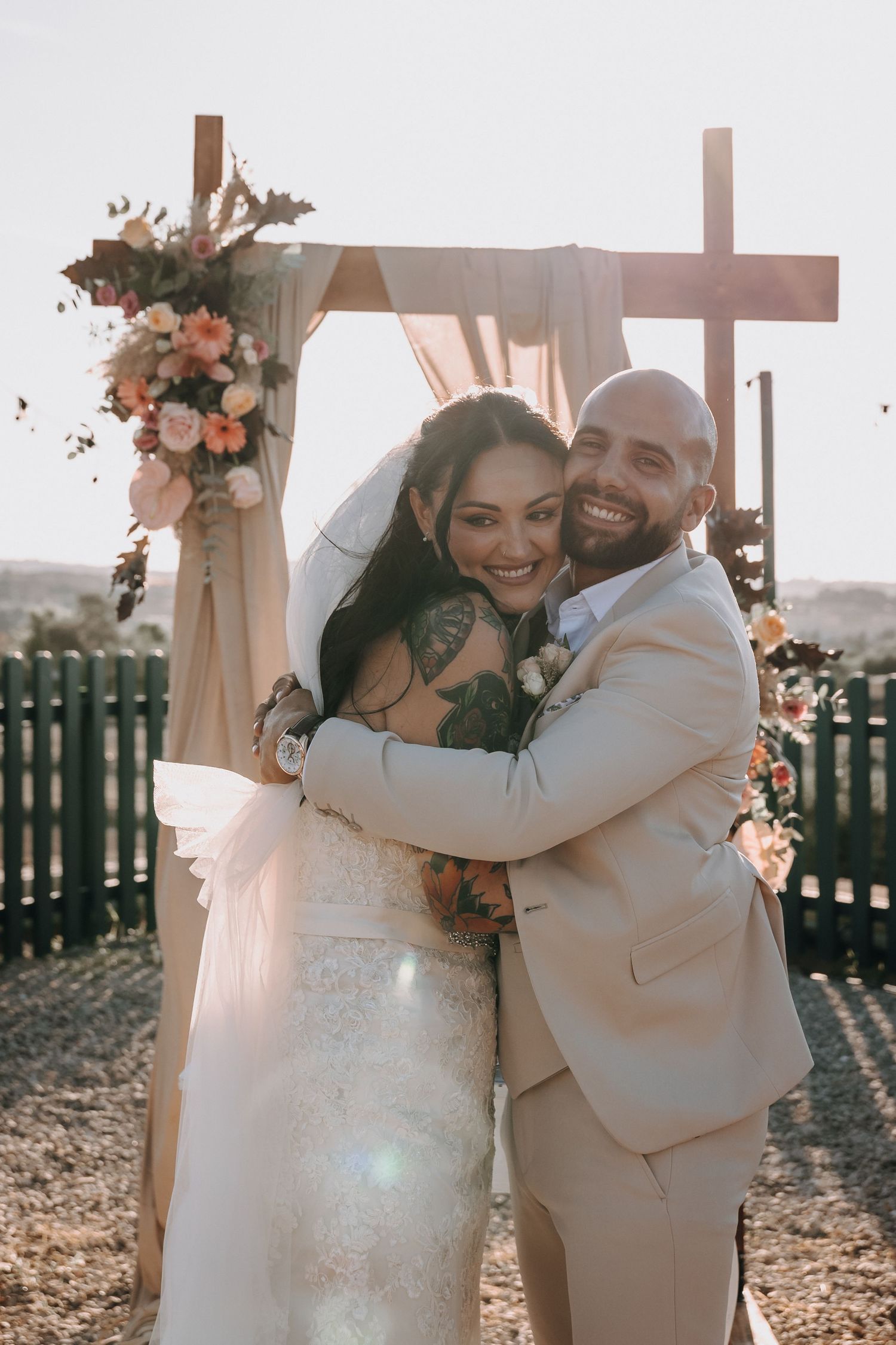 A happy couple embraces outdoors at sunset with a wooden cross and floral decorations in the background.