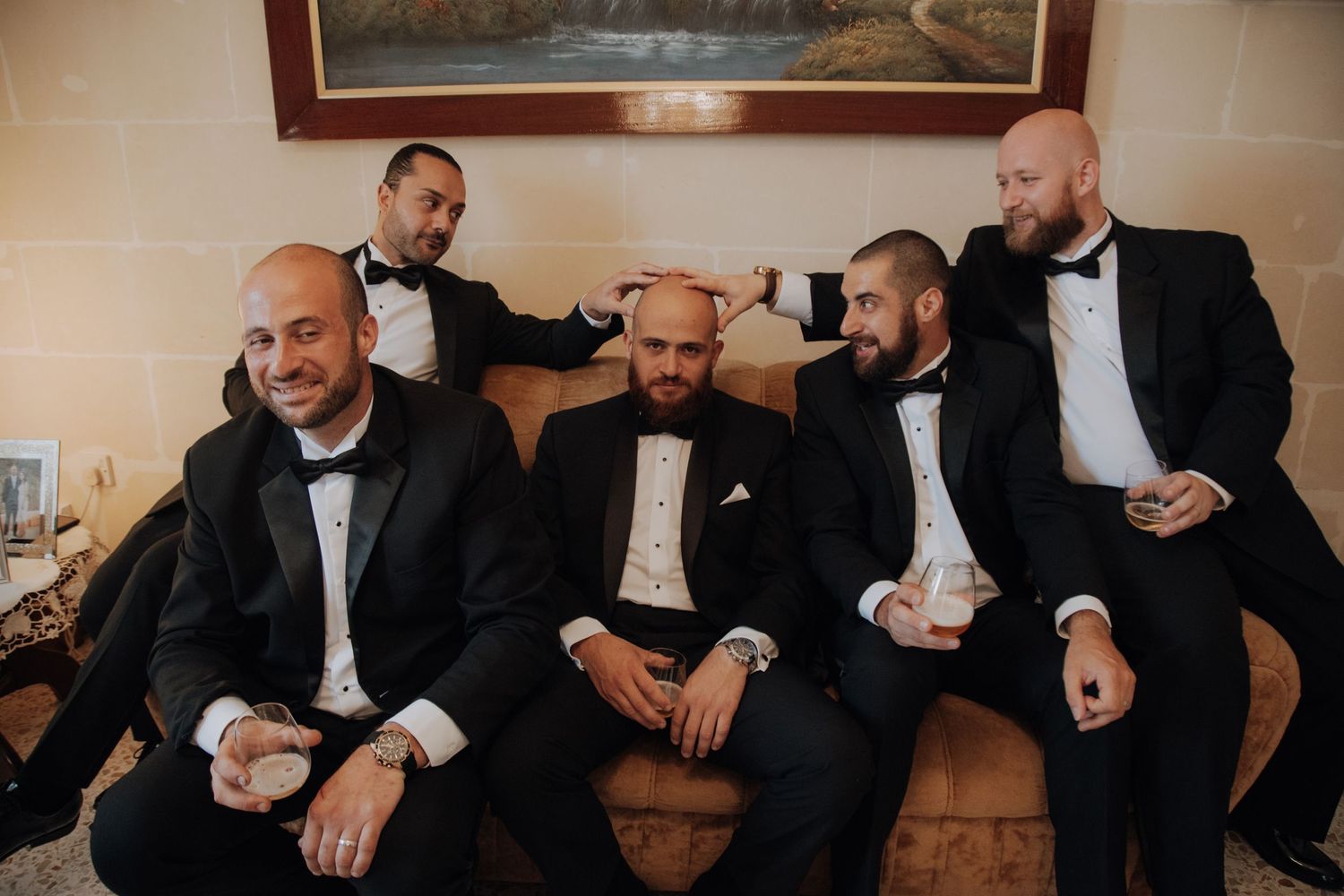 Several men in formal attire gather for drinks at an event with framed artwork in the background.
