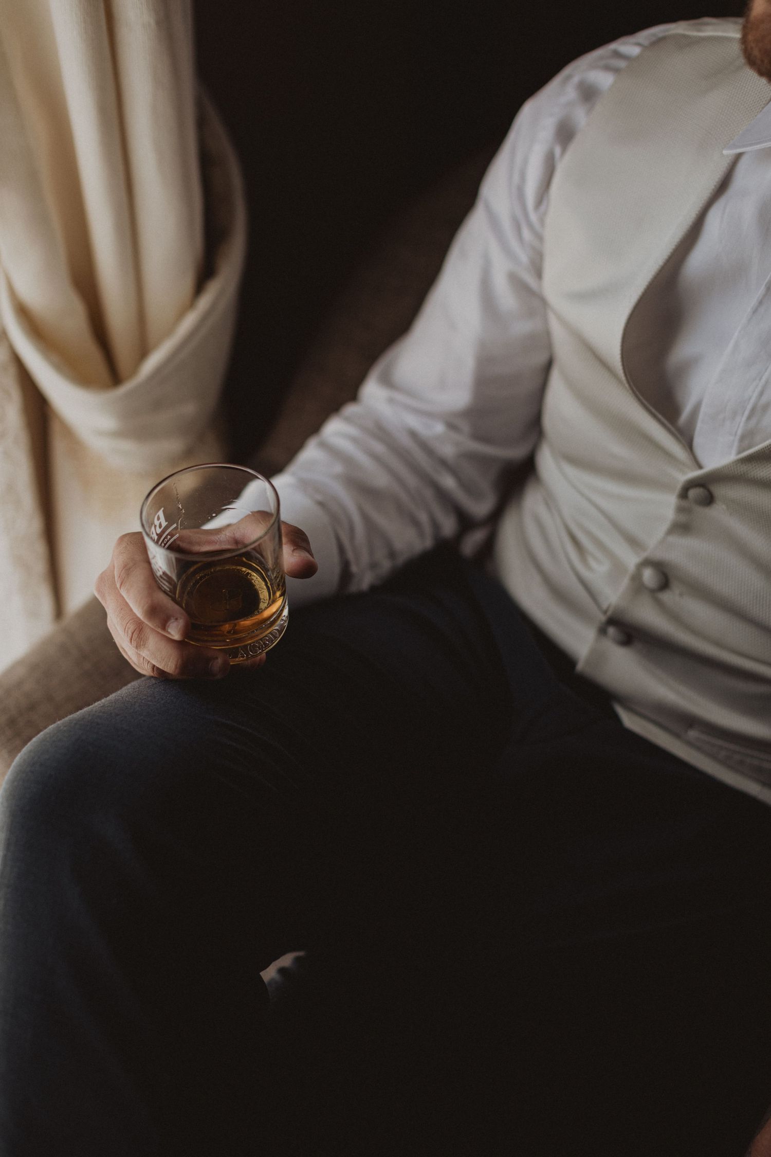 Person in white shirt holding a glass of whiskey while sitting.