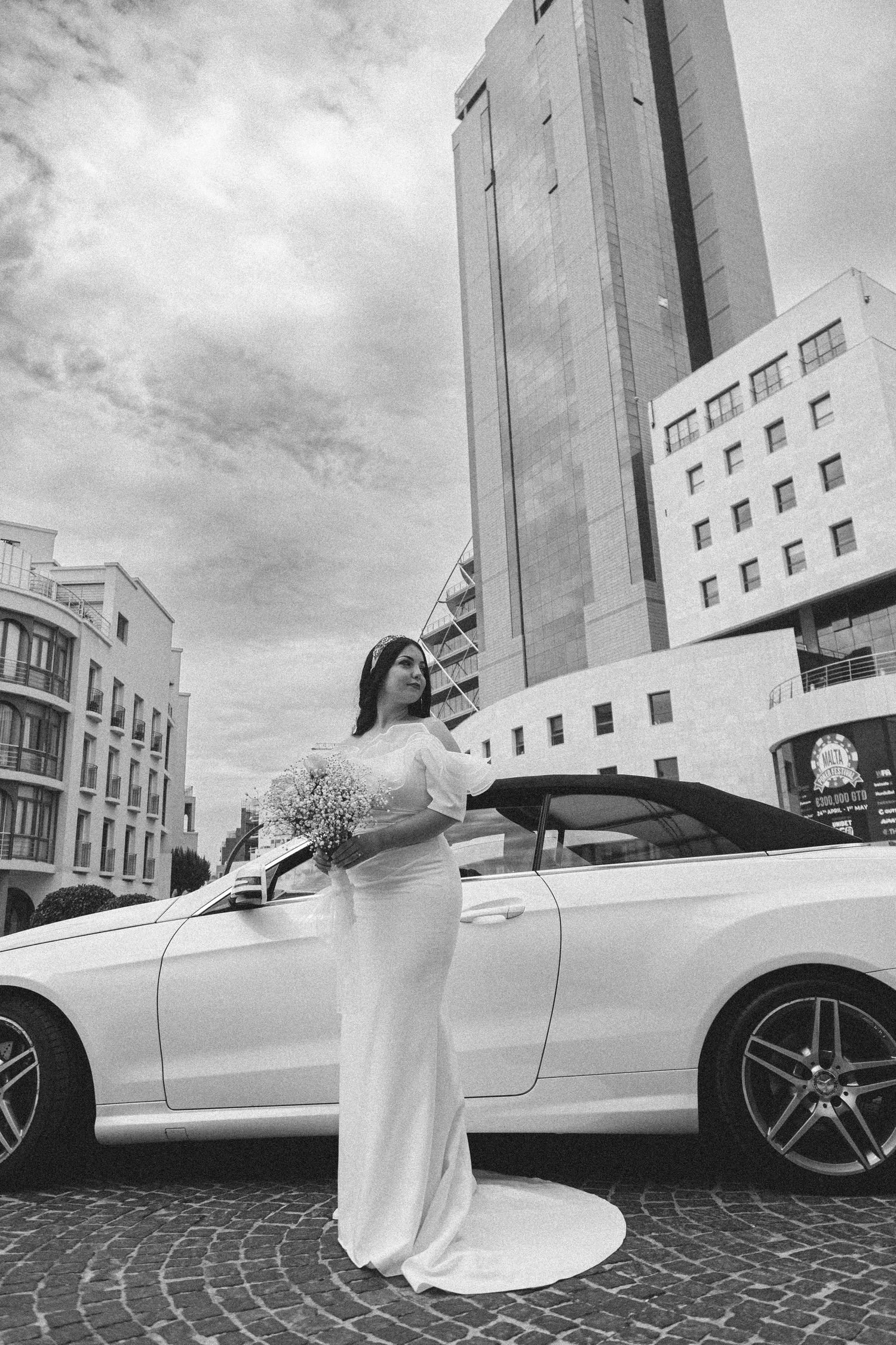 Elegant bridal portrait with white Mercedes and modern city architecture in background.