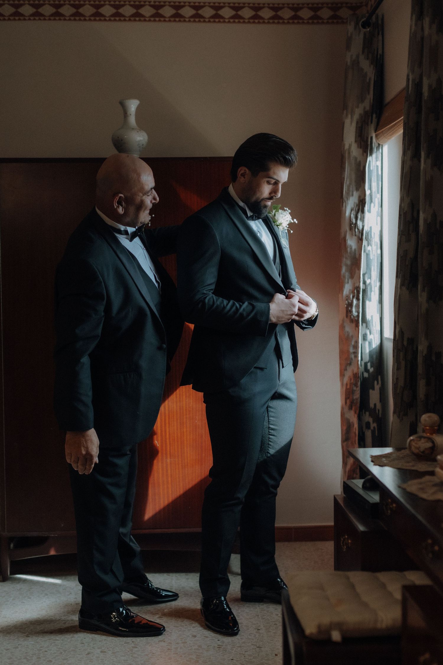 Two men in tuxedos standing by window in formal preparation room.