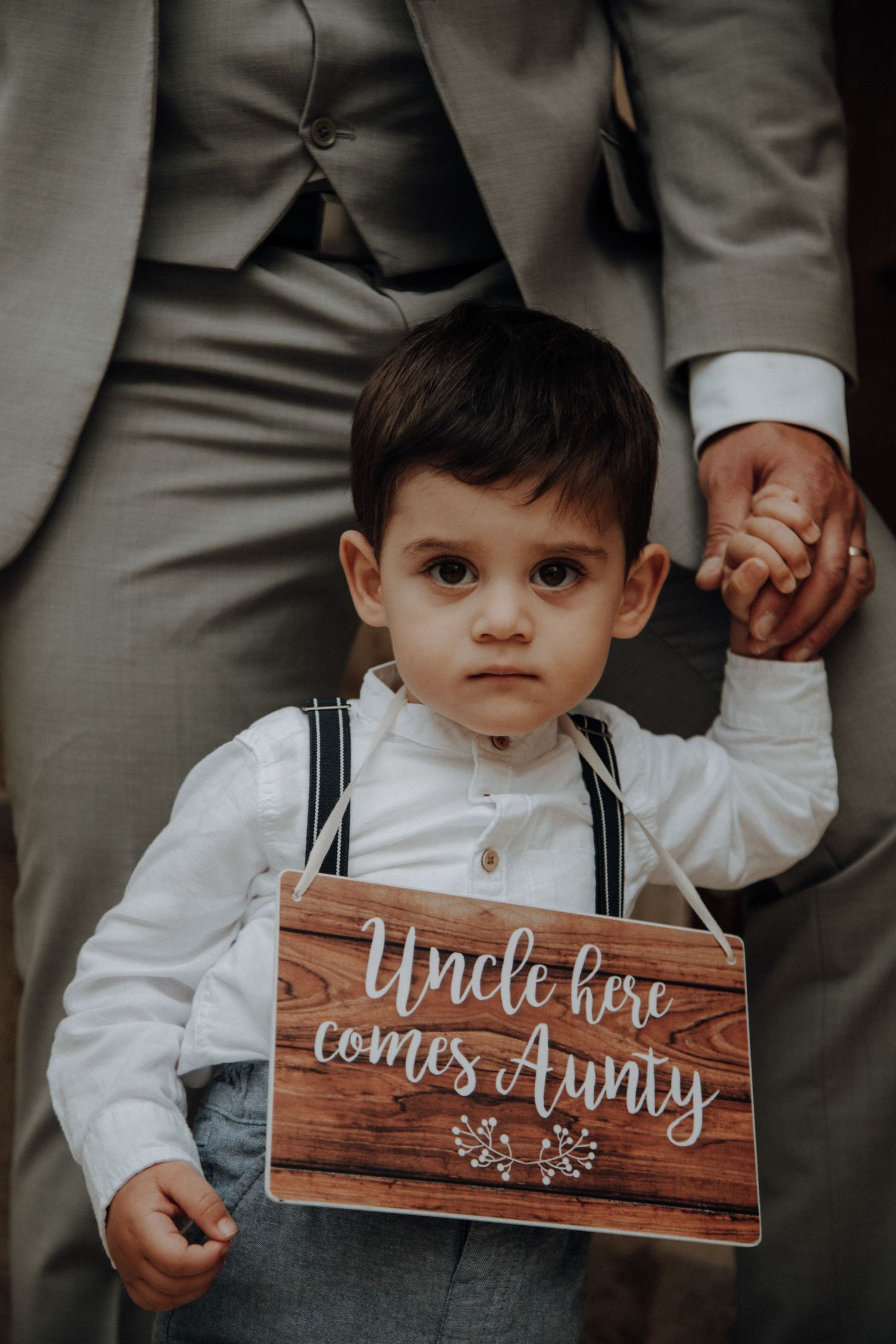 Small child in suspenders holds wooden wedding sign with text.