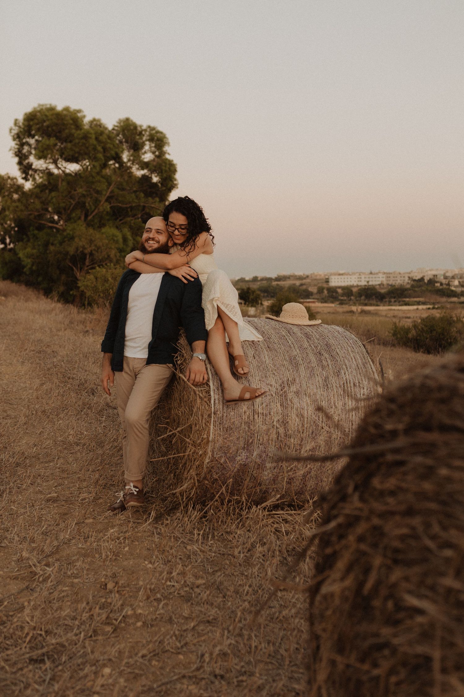 A playful moment captured between a couple on a hay bale during sunset with countryside views.