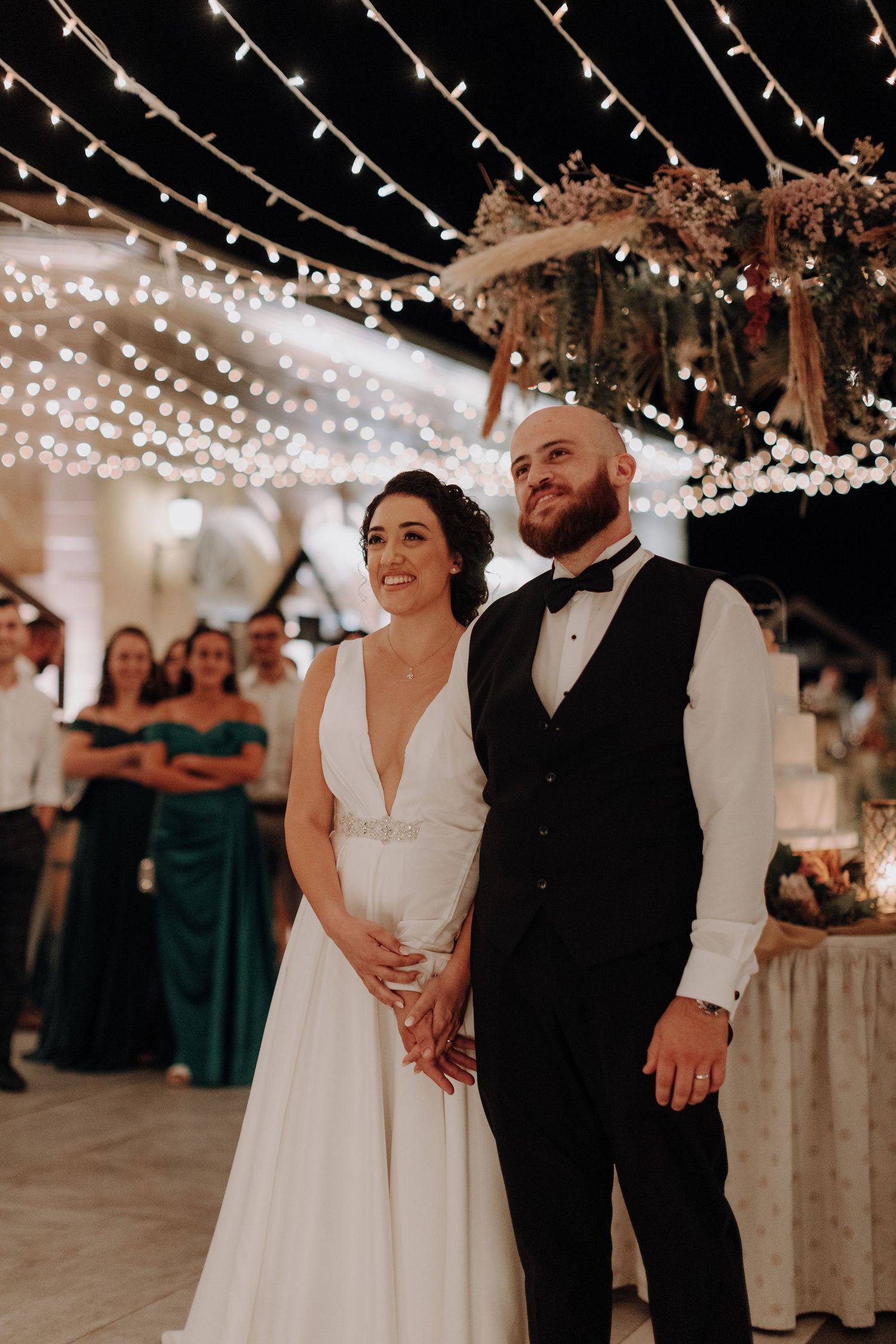 A newlywed couple stands under twinkling string lights at their evening reception.