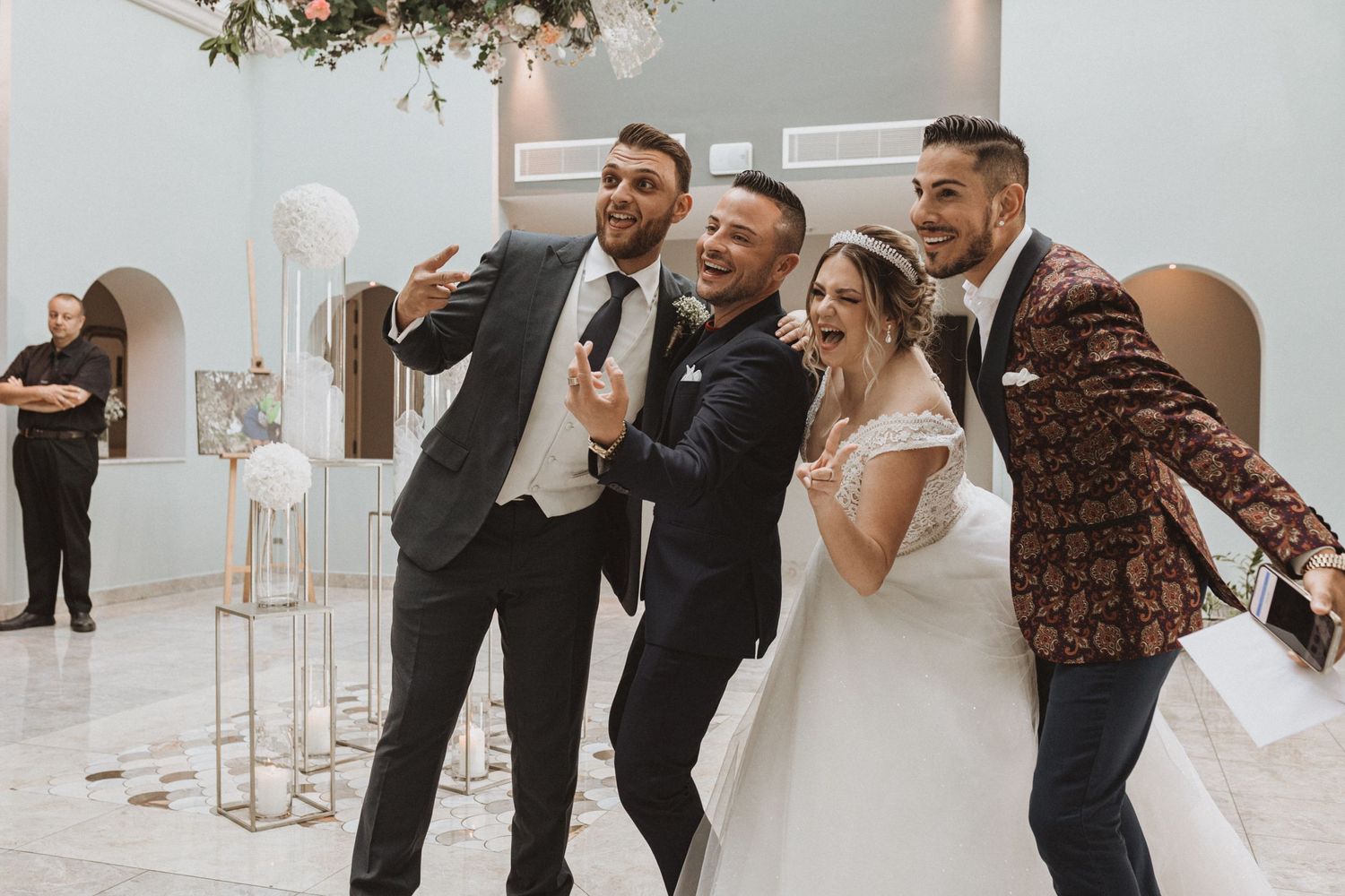 Wedding party members pose playfully for photos in elegant white reception venue.