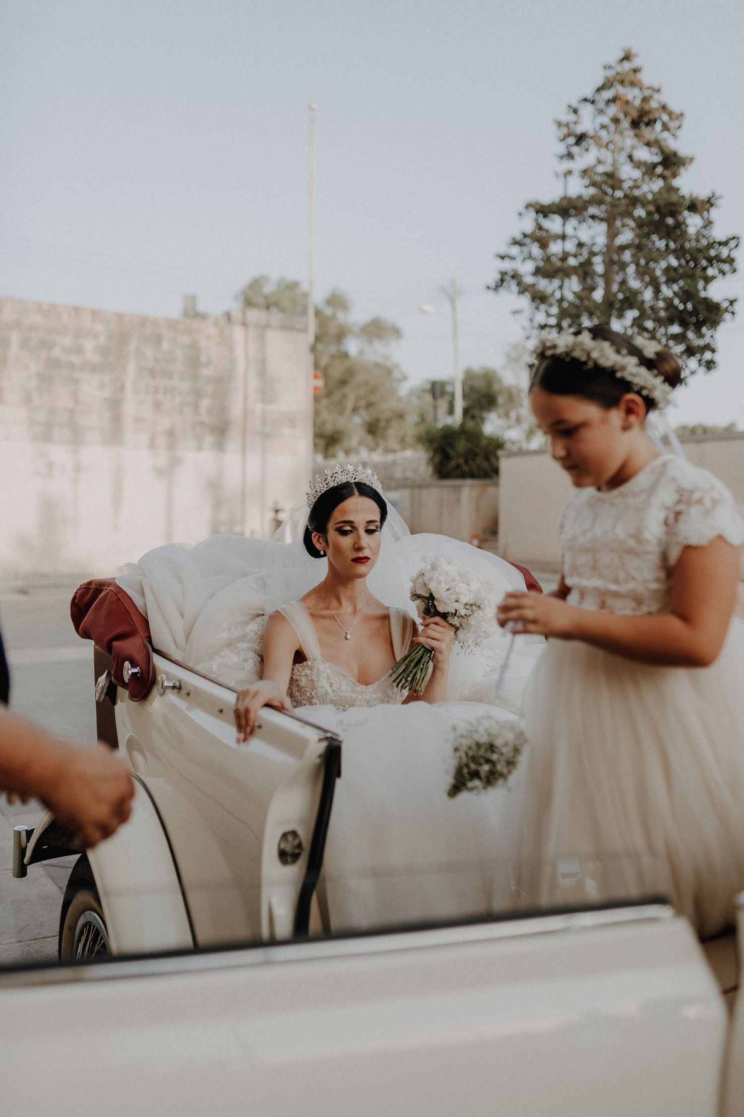 Bride in vintage car wearing crystal crown and lace gown during outdoor wedding.