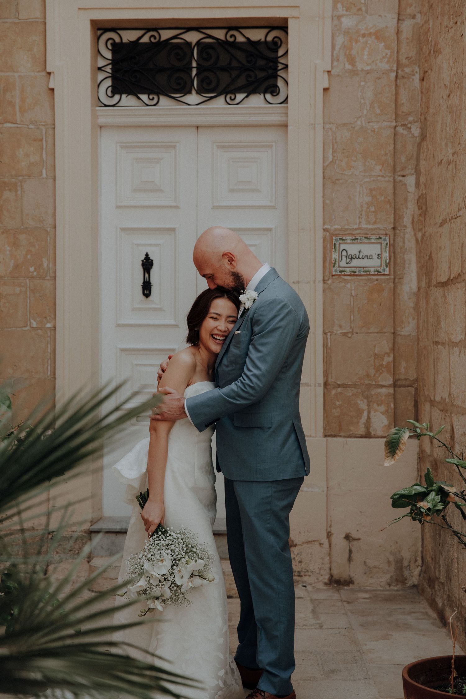 A tender wedding moment captured against a white door and stone wall with delicate palm fronds in the foreground.