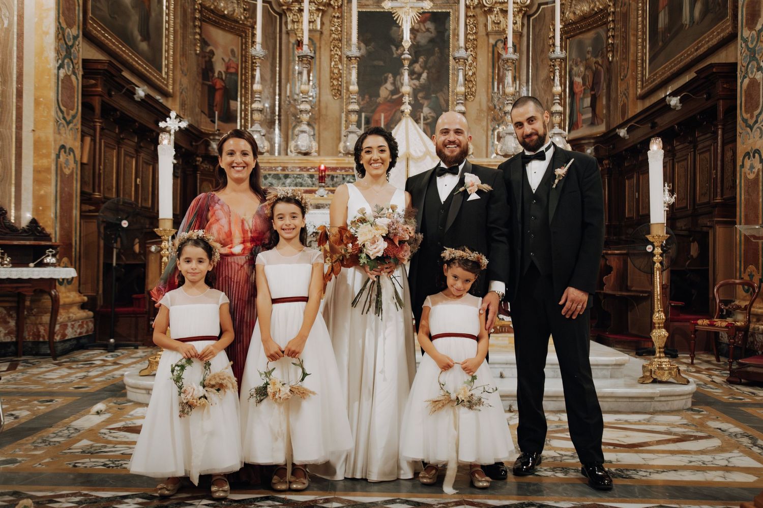 Wedding party with flower girls in white dresses pose in front of ornate church altar.