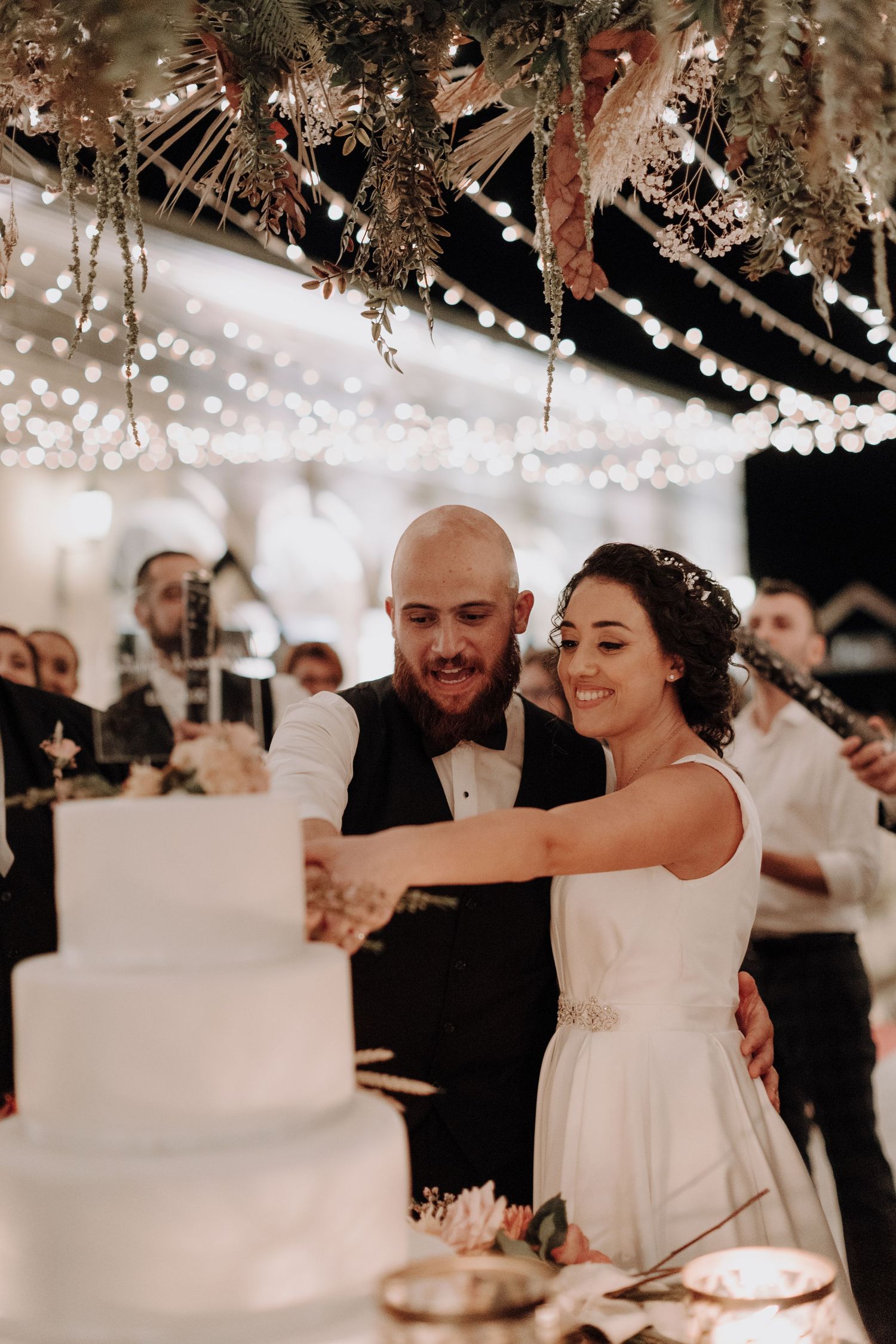 A couple cuts their elegant white wedding cake under sparkling fairy lights and dried floral decorations.