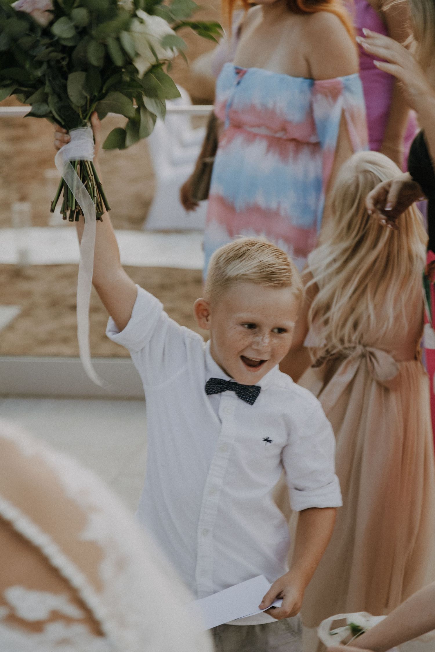 Young ring bearer in white shirt and bowtie joyfully holds up a bouquet.