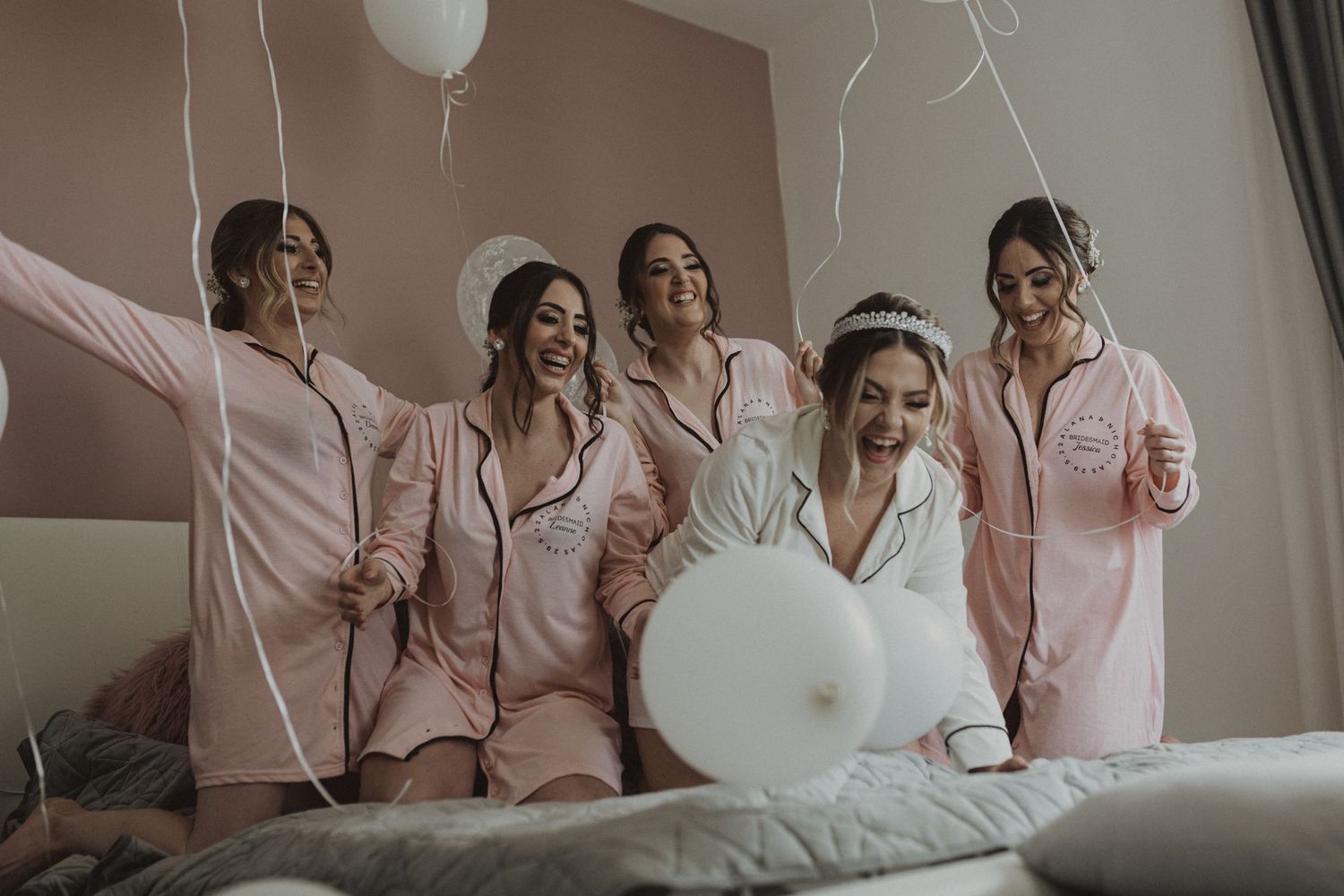 Candid moment of bridal party in pink robes holding white balloons in bedroom setting.