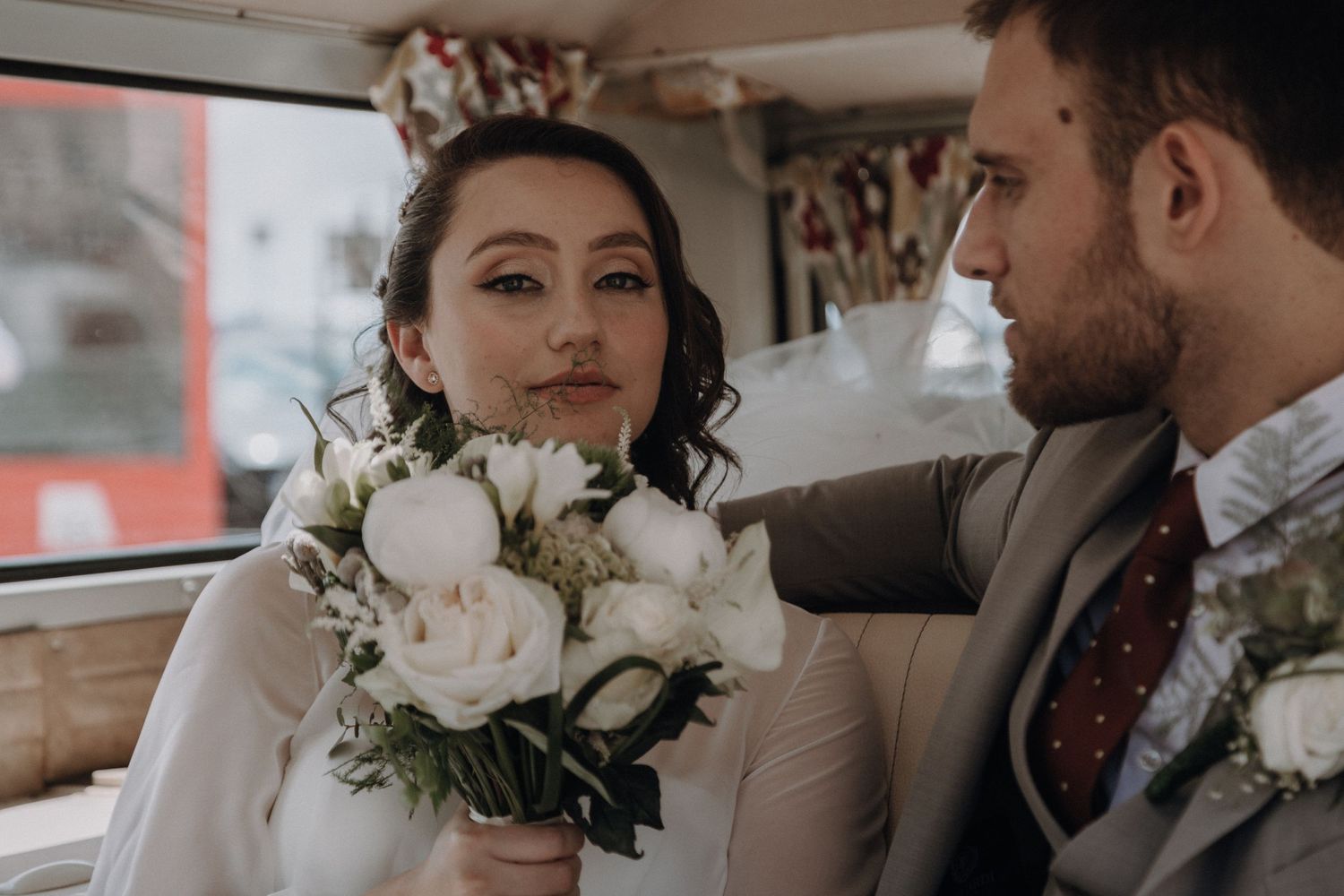 A bride holds a white rose bouquet while sitting next to her groom in a vintage vehicle.