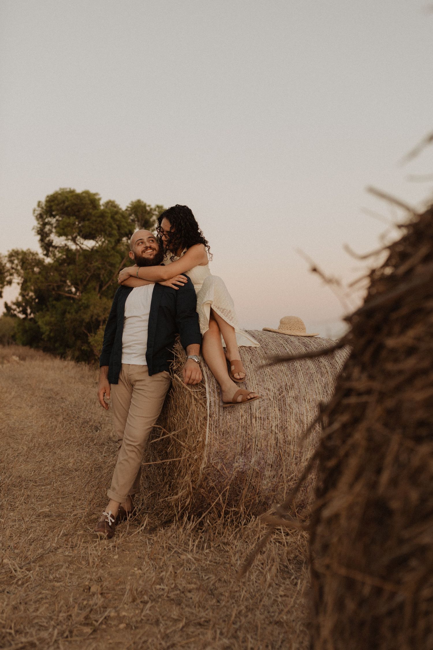 A couple shares a tender moment near hay bales during golden hour on a scenic hilltop.