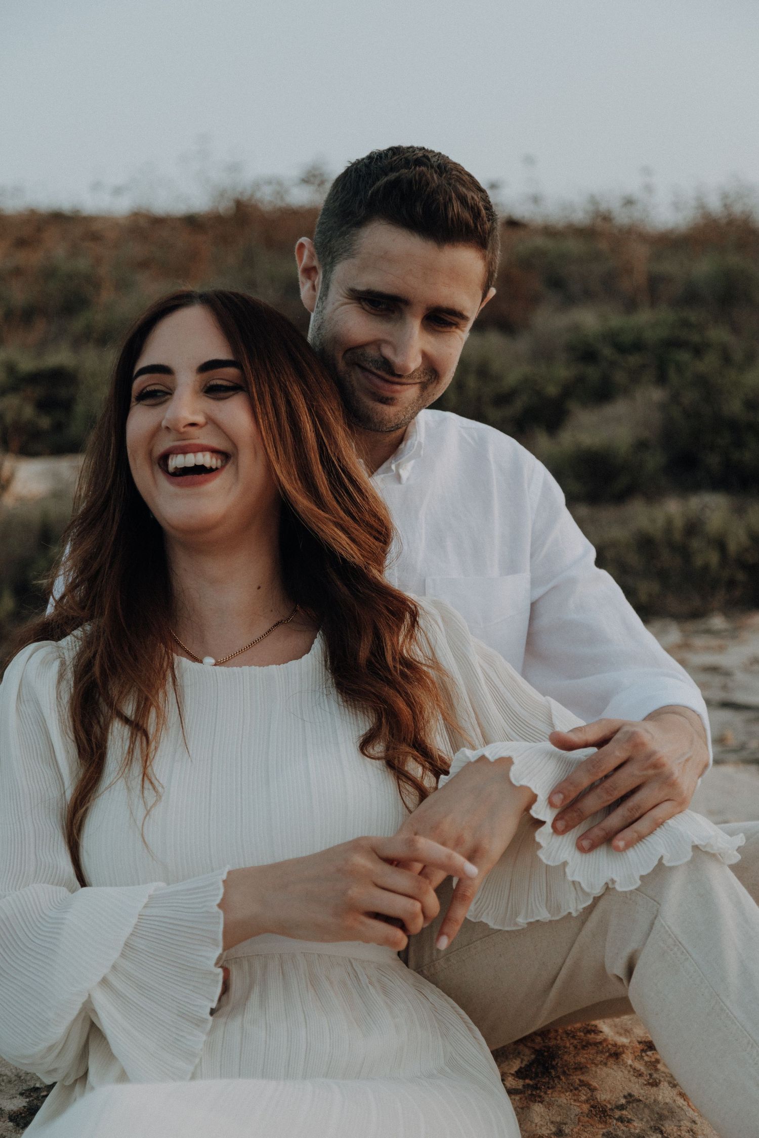 Romantic couple in white outfits embracing and laughing during a coastal photoshoot.
