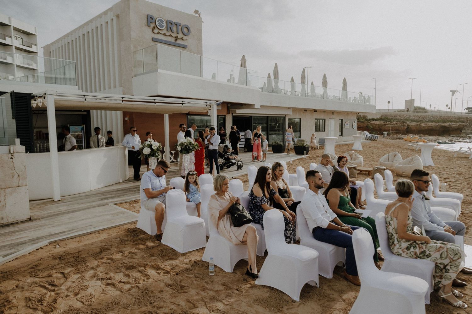 Wedding guests seated in white chairs outside Porto restaurant for a beach ceremony.