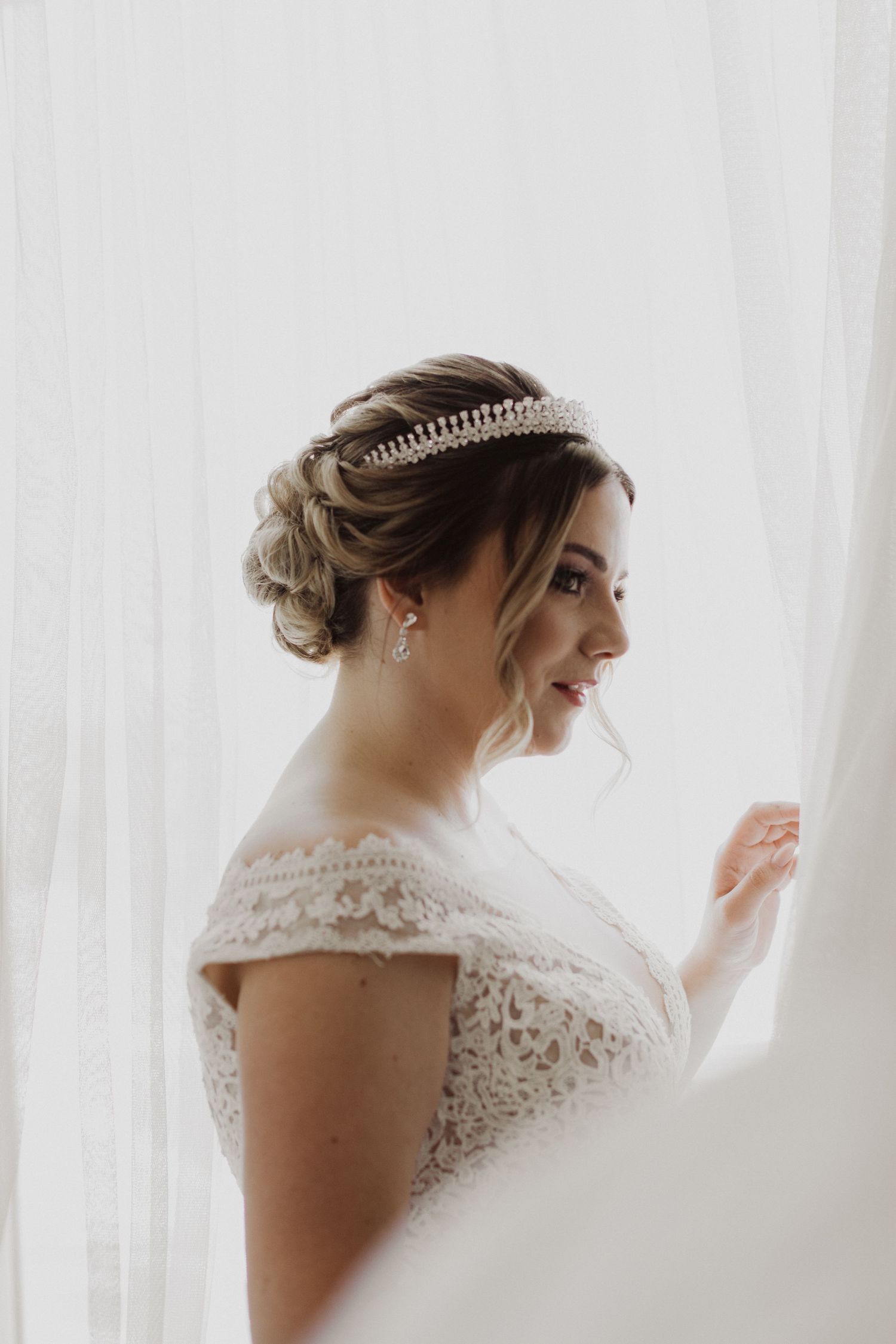 Side profile of a bride in an off-shoulder lace gown and pearl tiara near a window.