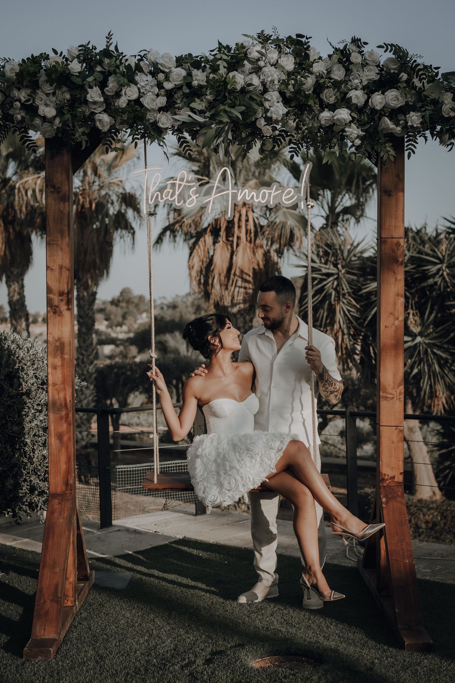 A couple shares a romantic moment on a swing under a floral arch with palm trees in the background.