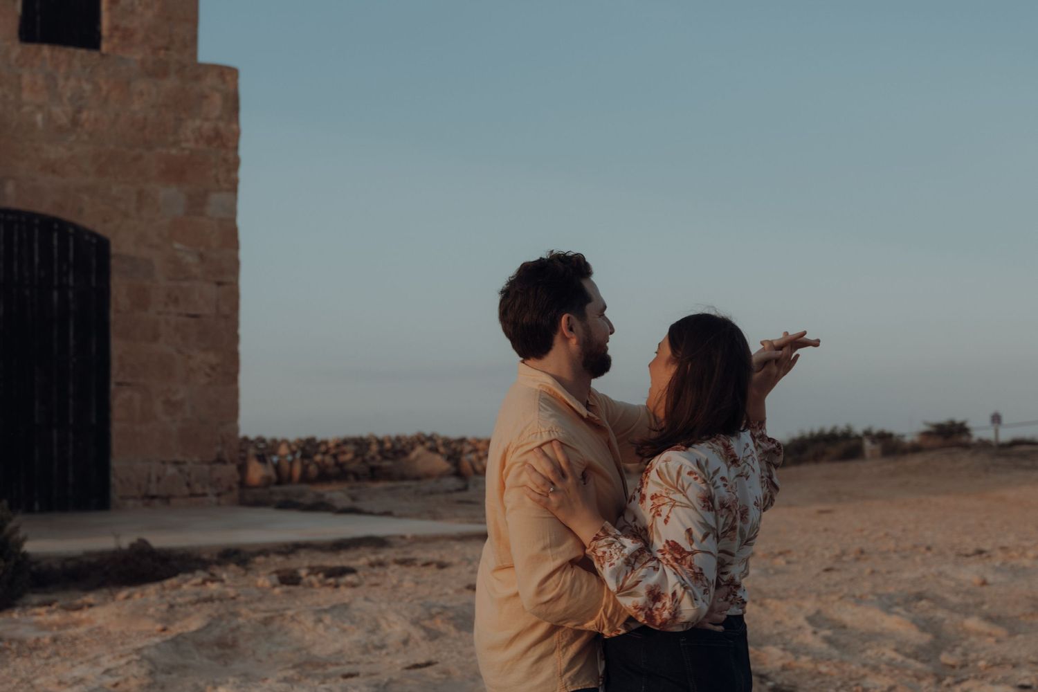 A couple stands together in a desert landscape during golden hour with stone architecture in the background.