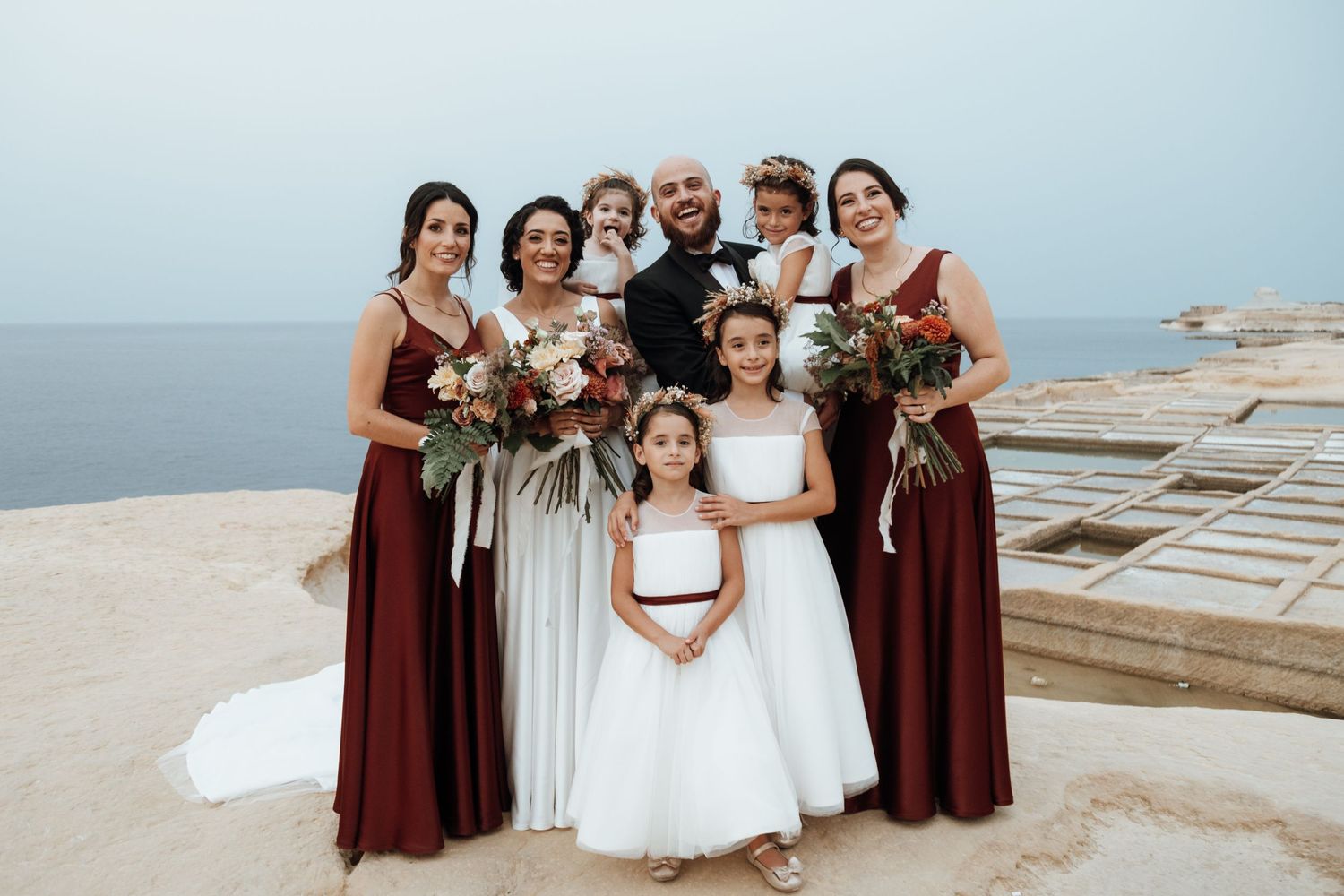 A bridal party in burgundy dresses gathers for a photo by the ocean.