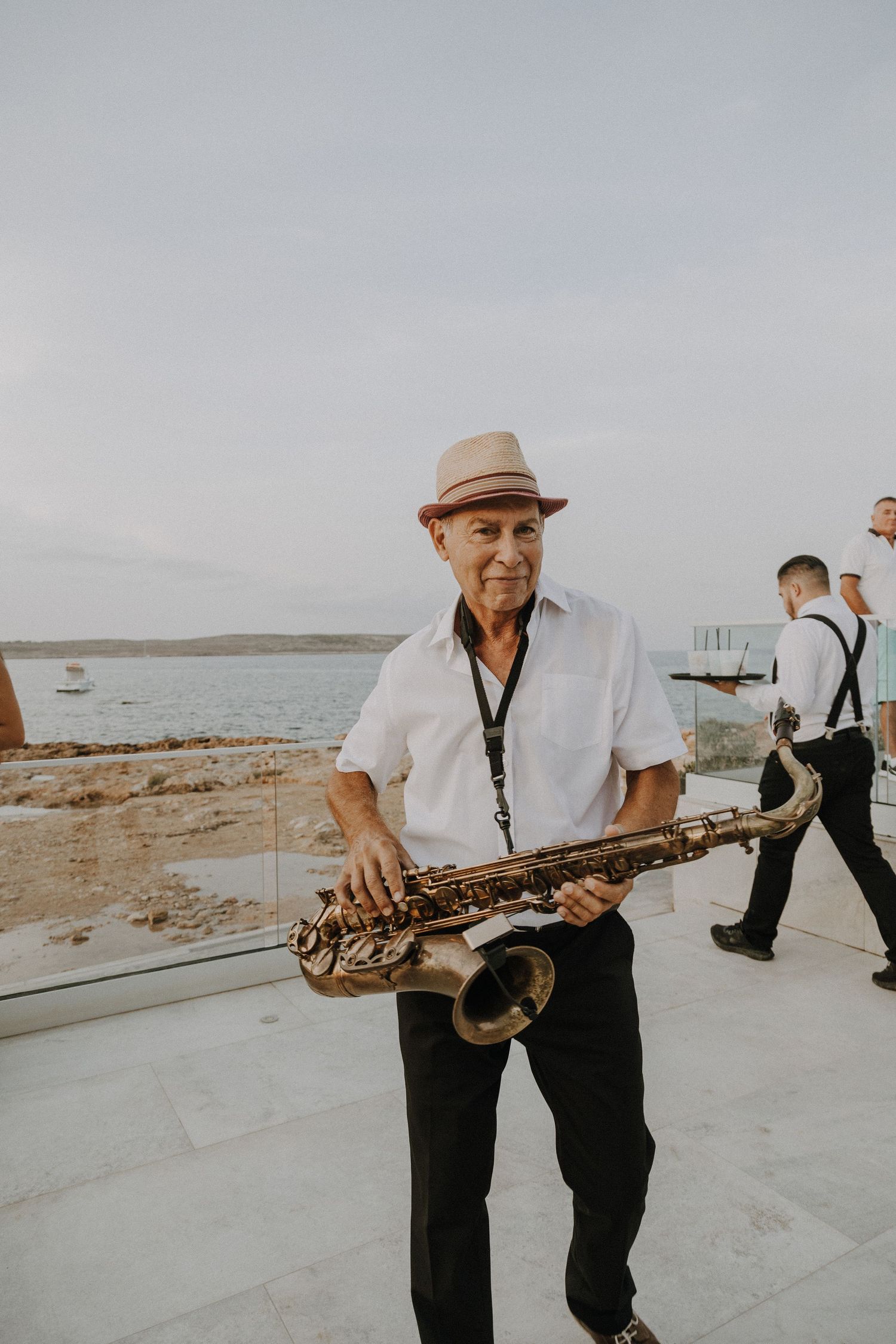 Musician in fedora hat plays saxophone at beachside wedding venue.