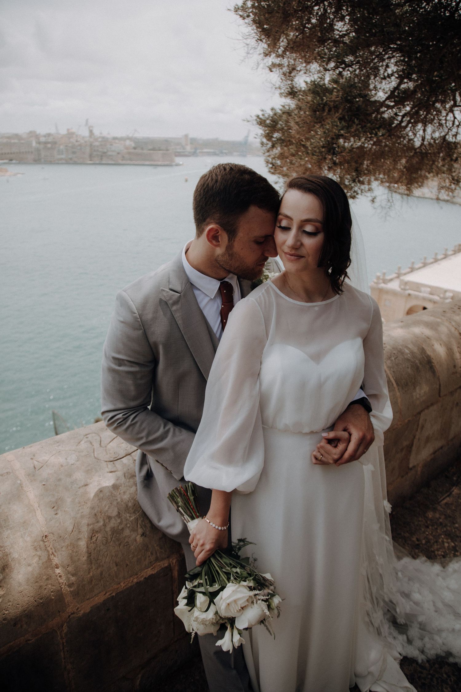 Wedding couple embracing by waterfront with bride in flowing white dress holding bouquet.