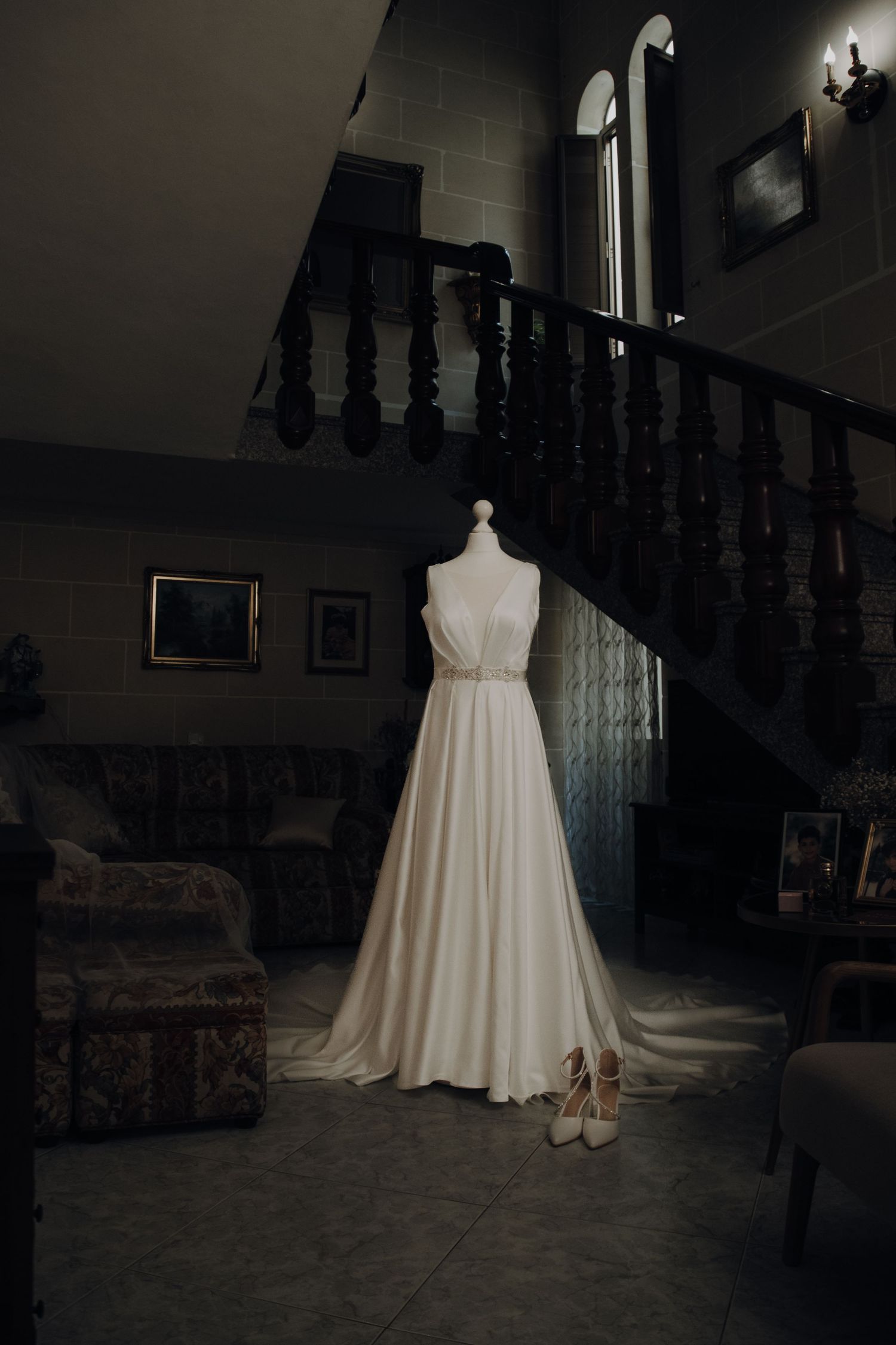 White wedding dress and shoes displayed on mannequin near dark wooden staircase.