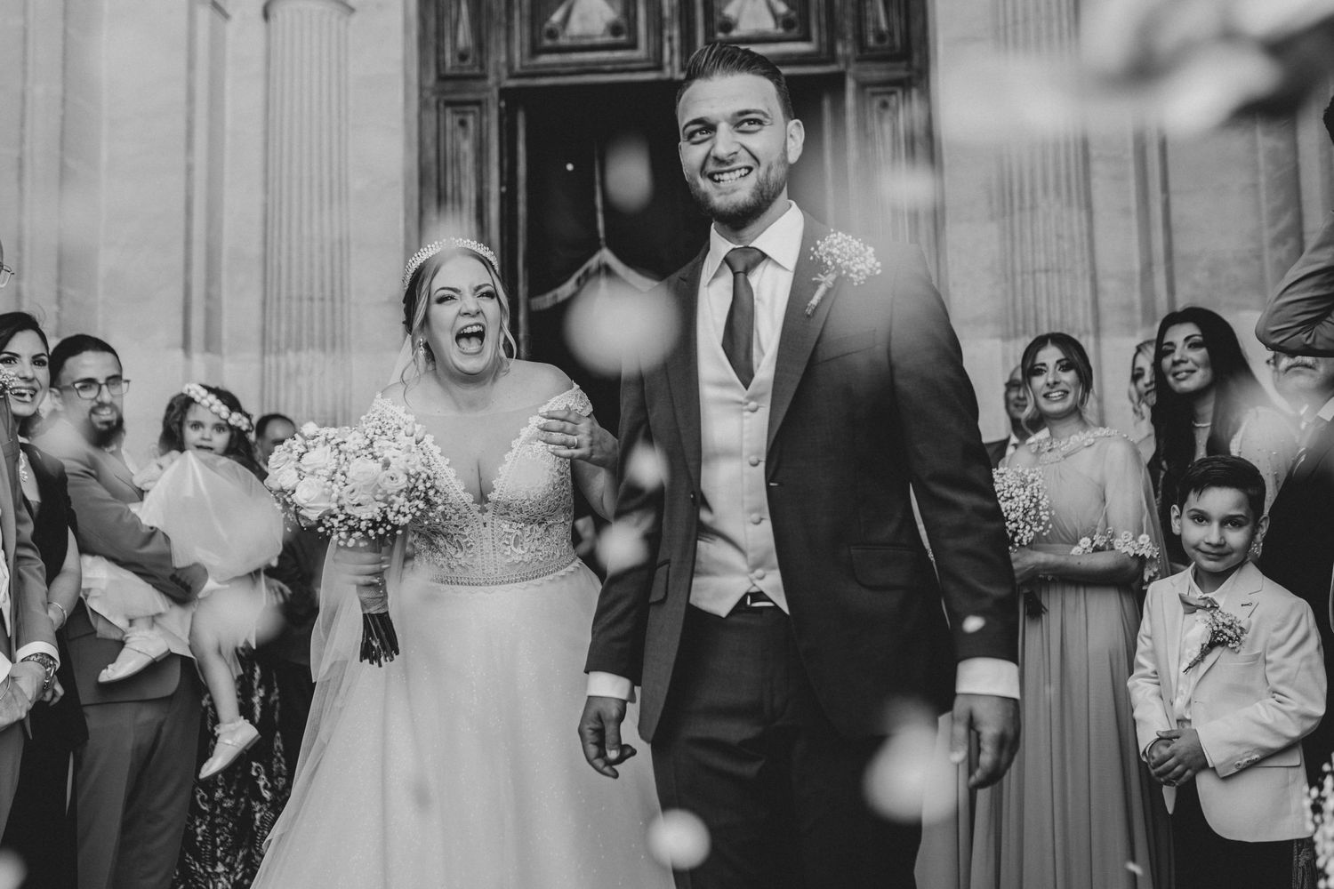 Black and white wedding photo capturing a joyful moment on steps outside a grand building with guests celebrating.