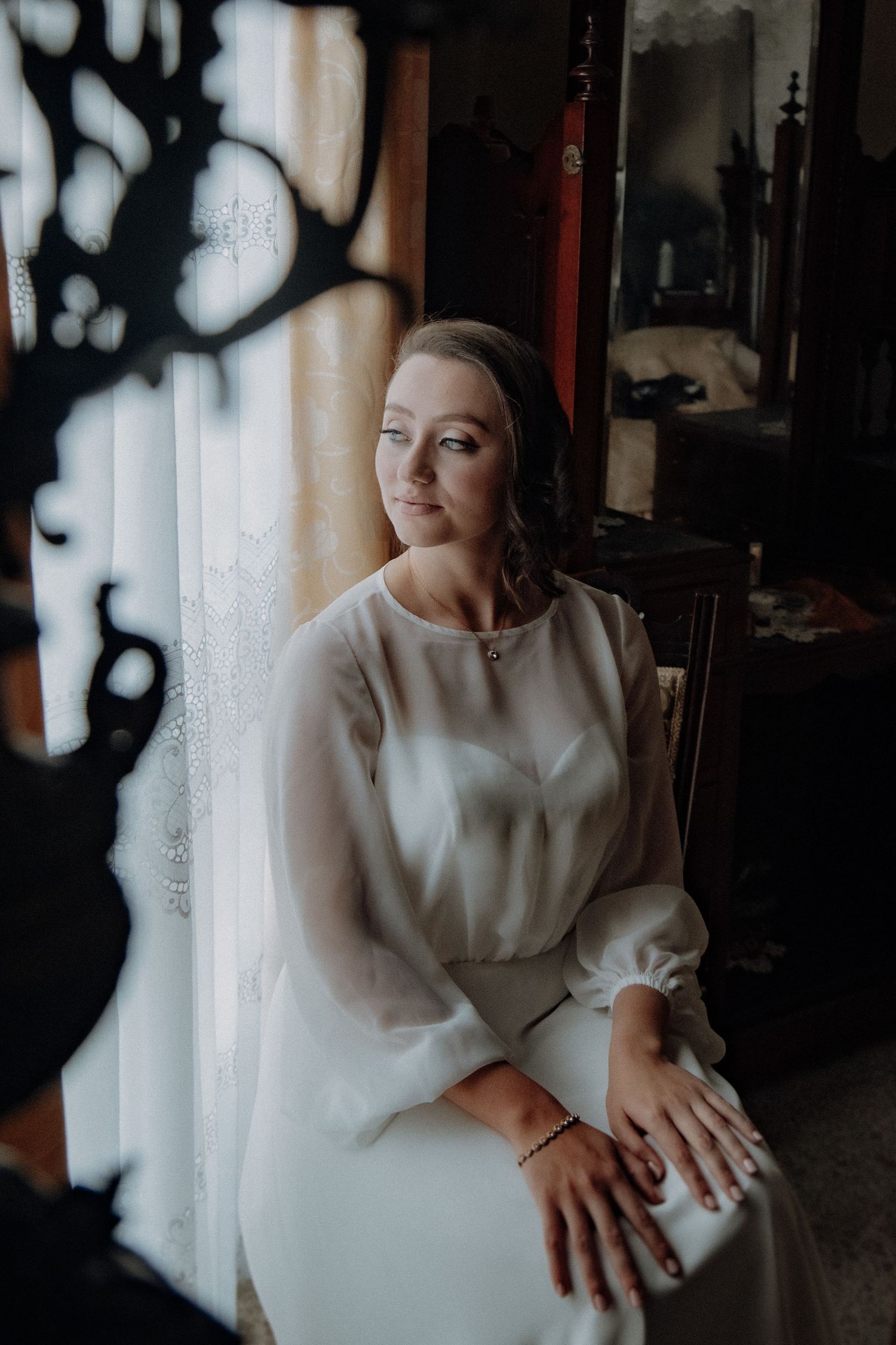 Moody window portrait of bride in flowing white dress with bishop sleeves.