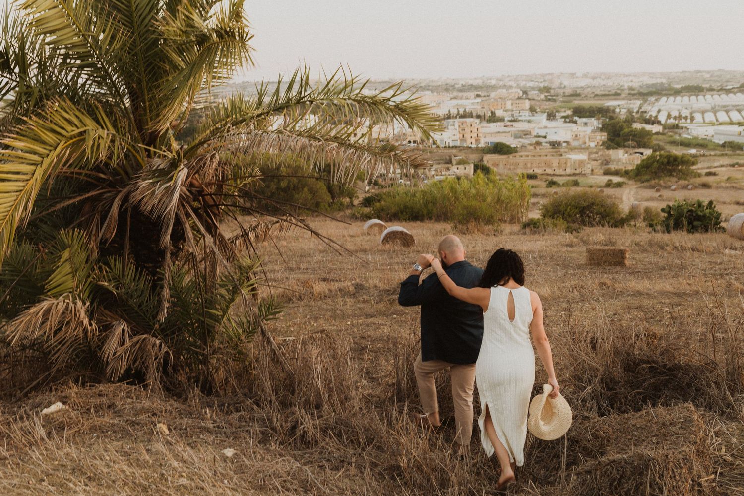 A couple walking down a hillside path with palm trees and a distant cityscape during golden hour.