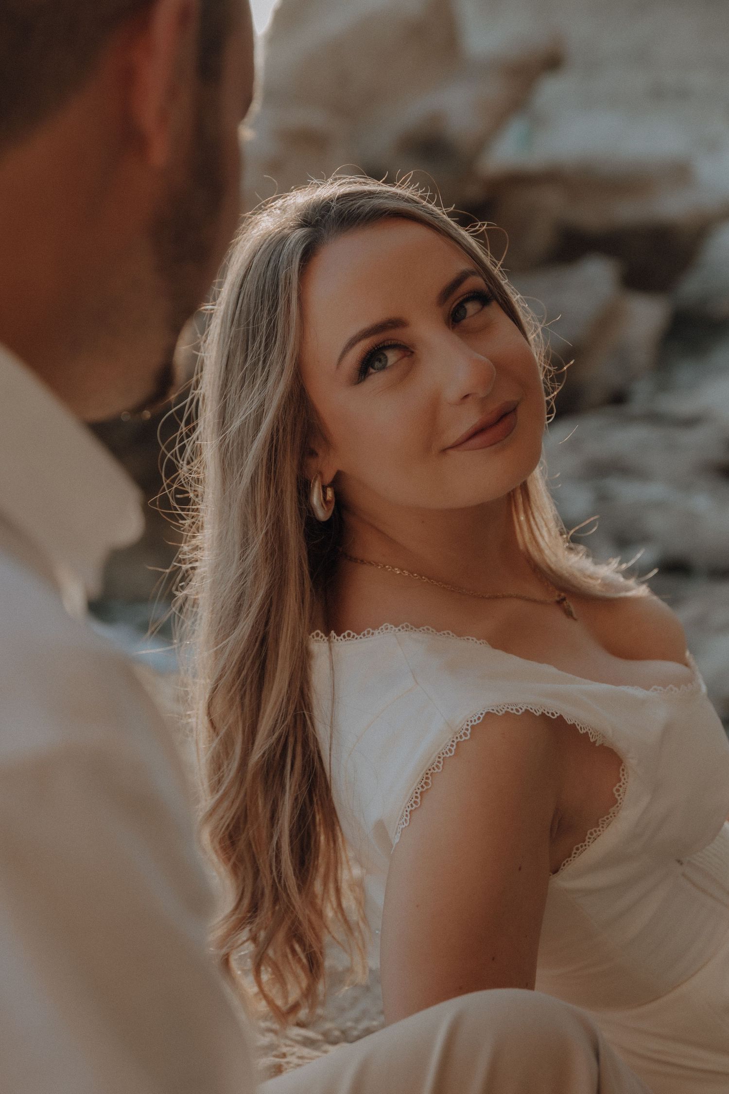A woman in a white off-shoulder dress gazes lovingly at her partner during a romantic outdoor engagement session.