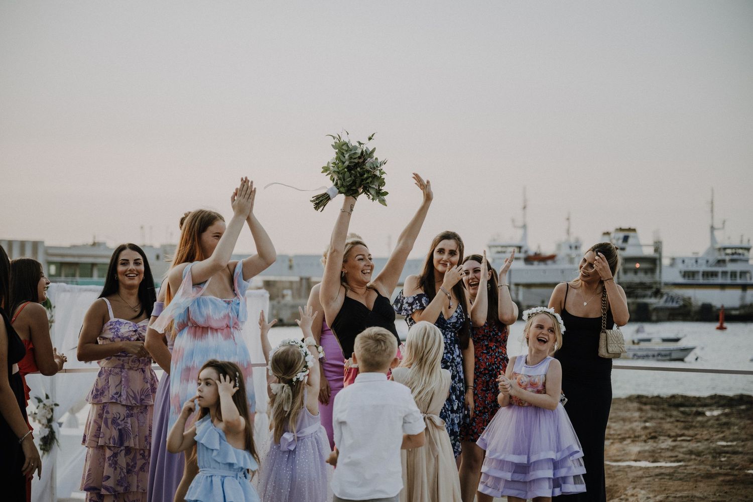 Wedding guests excitedly reach for the bouquet toss at a seaside venue with boats in the background.