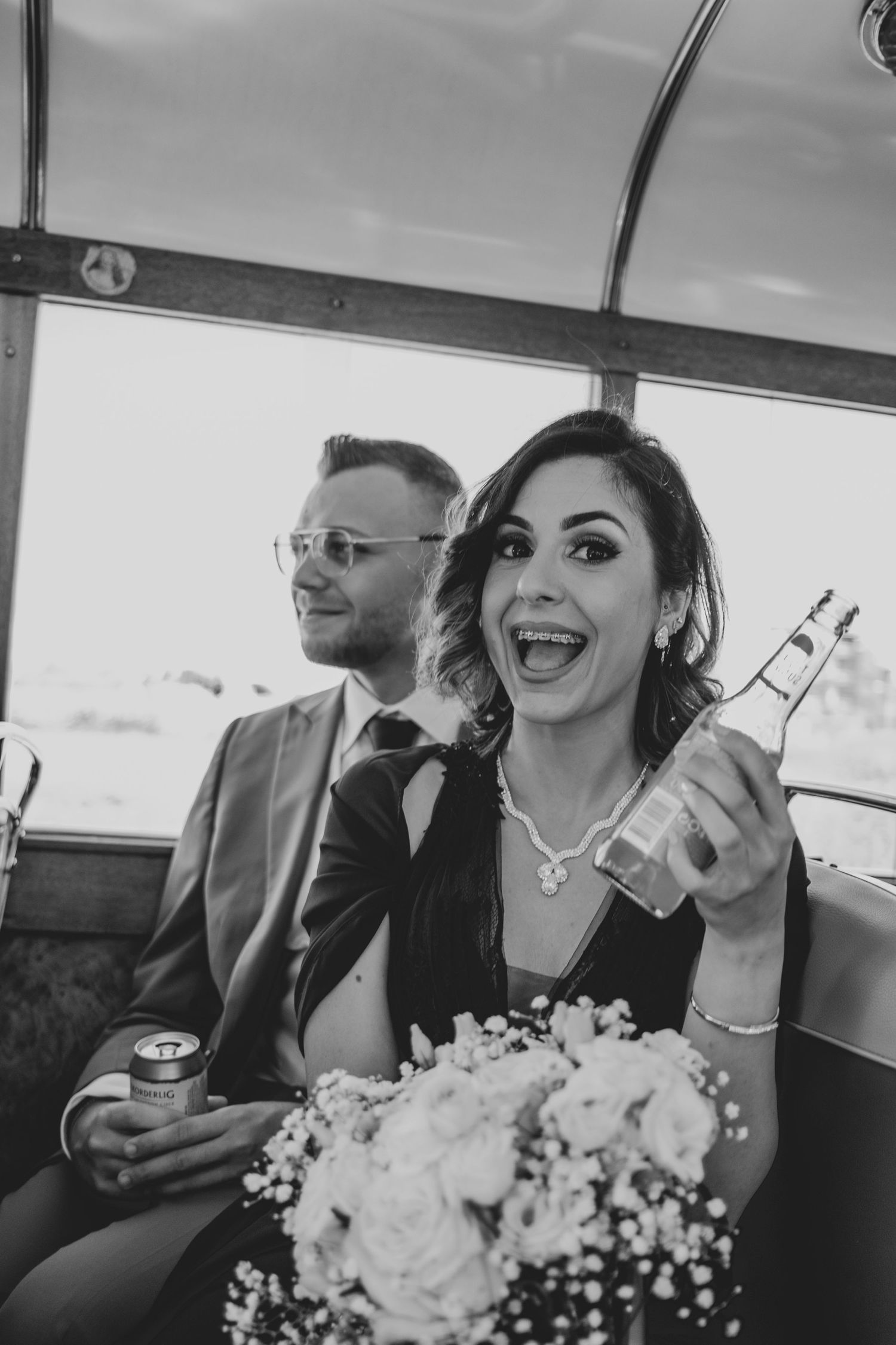 Bridesmaid in black dress laughs while holding beverage in vintage bus.