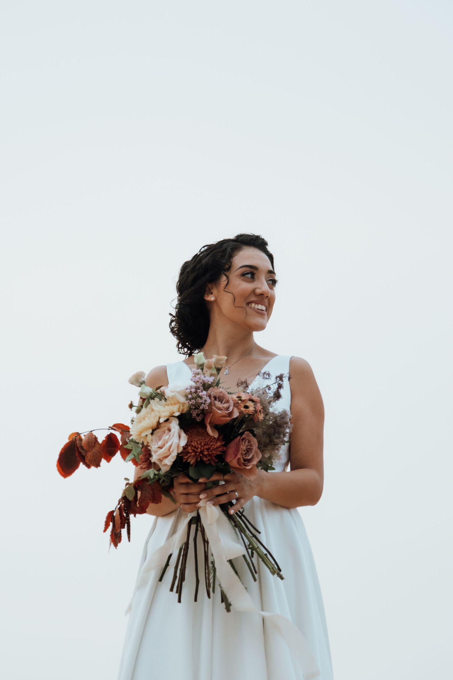 A radiant bride holding a beautiful fall-toned bouquet smiles against a cloudy coastal background.