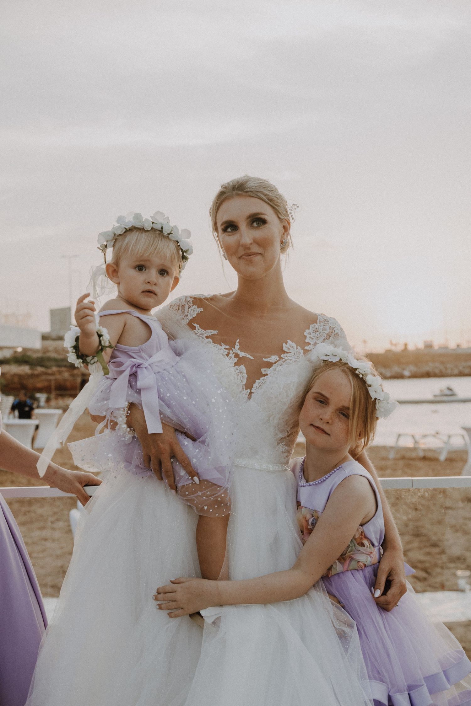 Elegant bride in white lace gown poses with young attendants in lavender dresses.