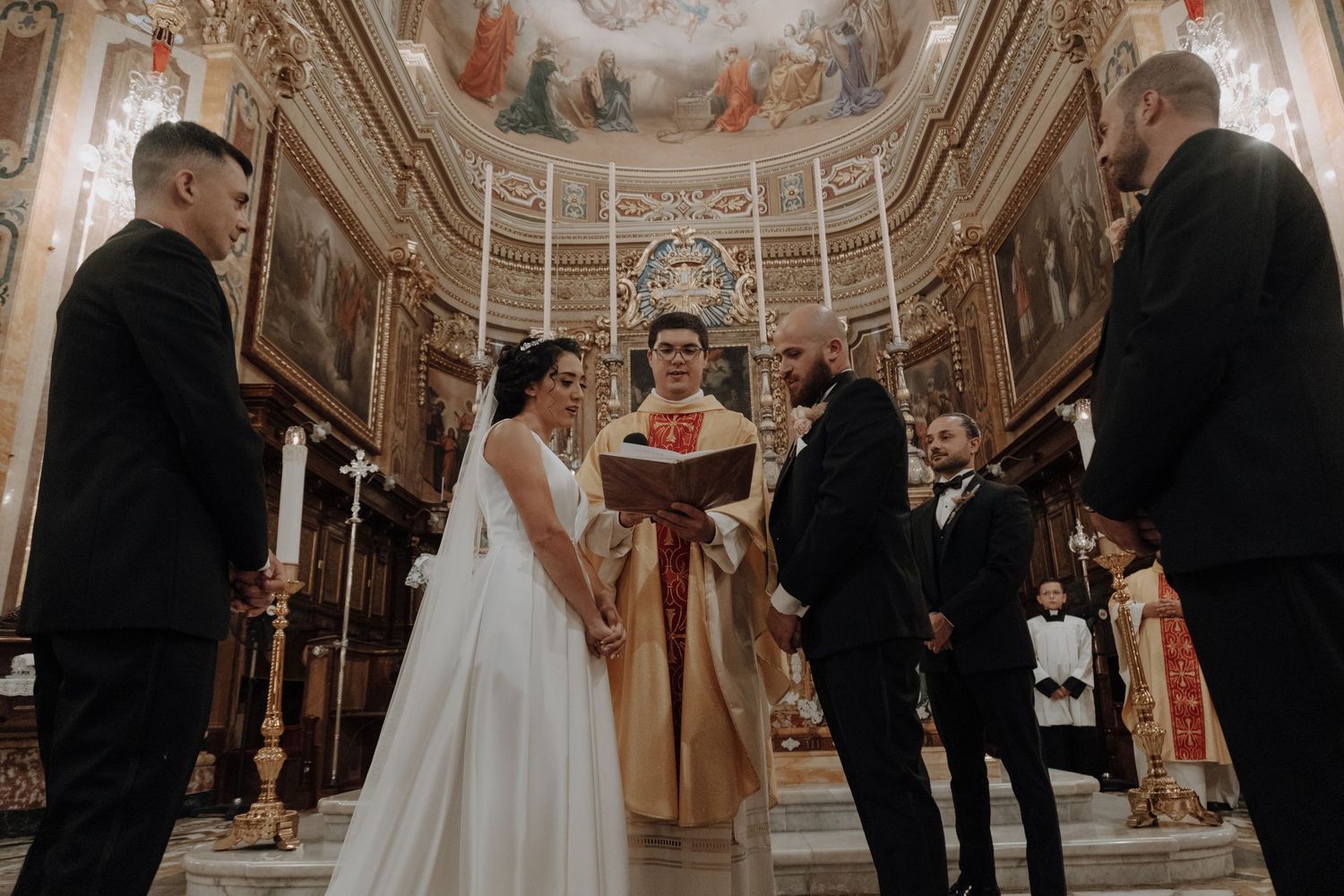 Wedding ceremony taking place in ornately decorated church with frescoed ceiling.