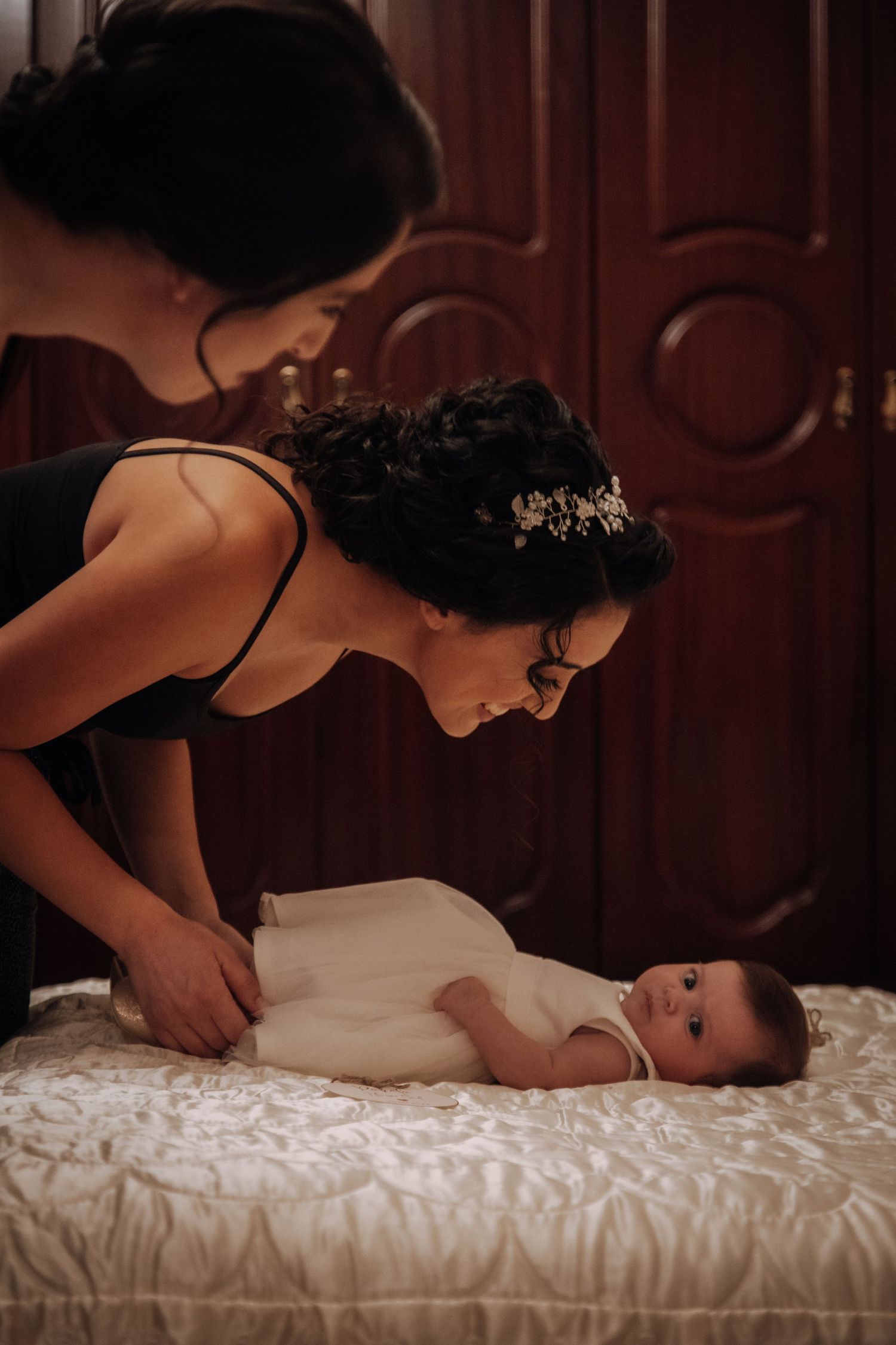 A person in dark attire leans over to interact with a baby lying on white bedding.