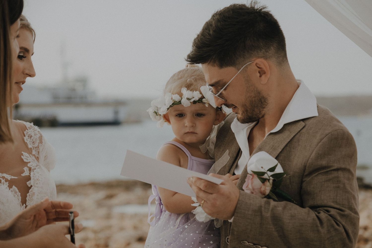 Wedding guest in tan suit holds paper while small child in white dress looks on.